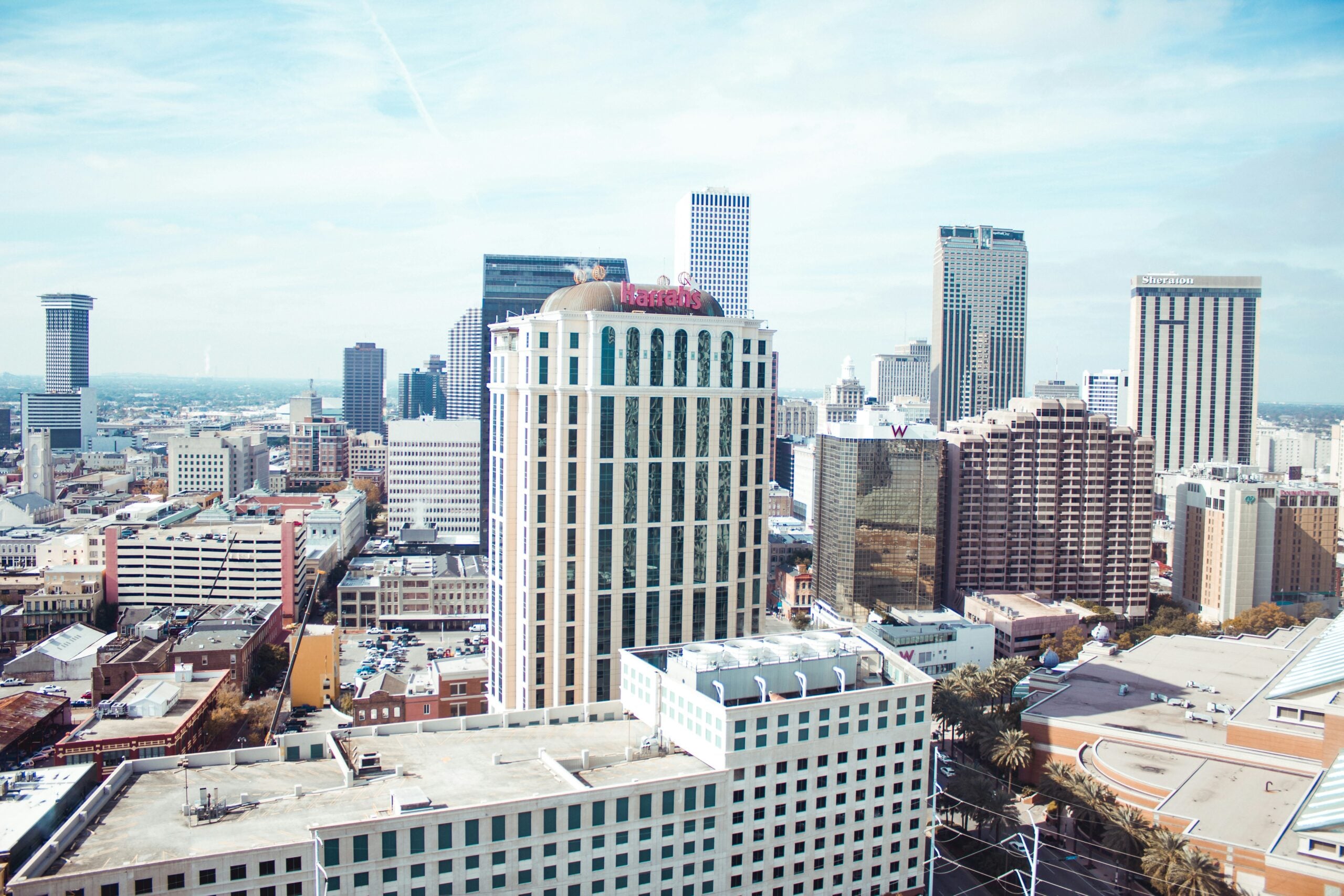 A bright cityscape of downtown New Orleans featuring tall office buildings, including the Harrah’s hotel prominently in the center, with a clear sky above and the Mississippi River in the distant background.