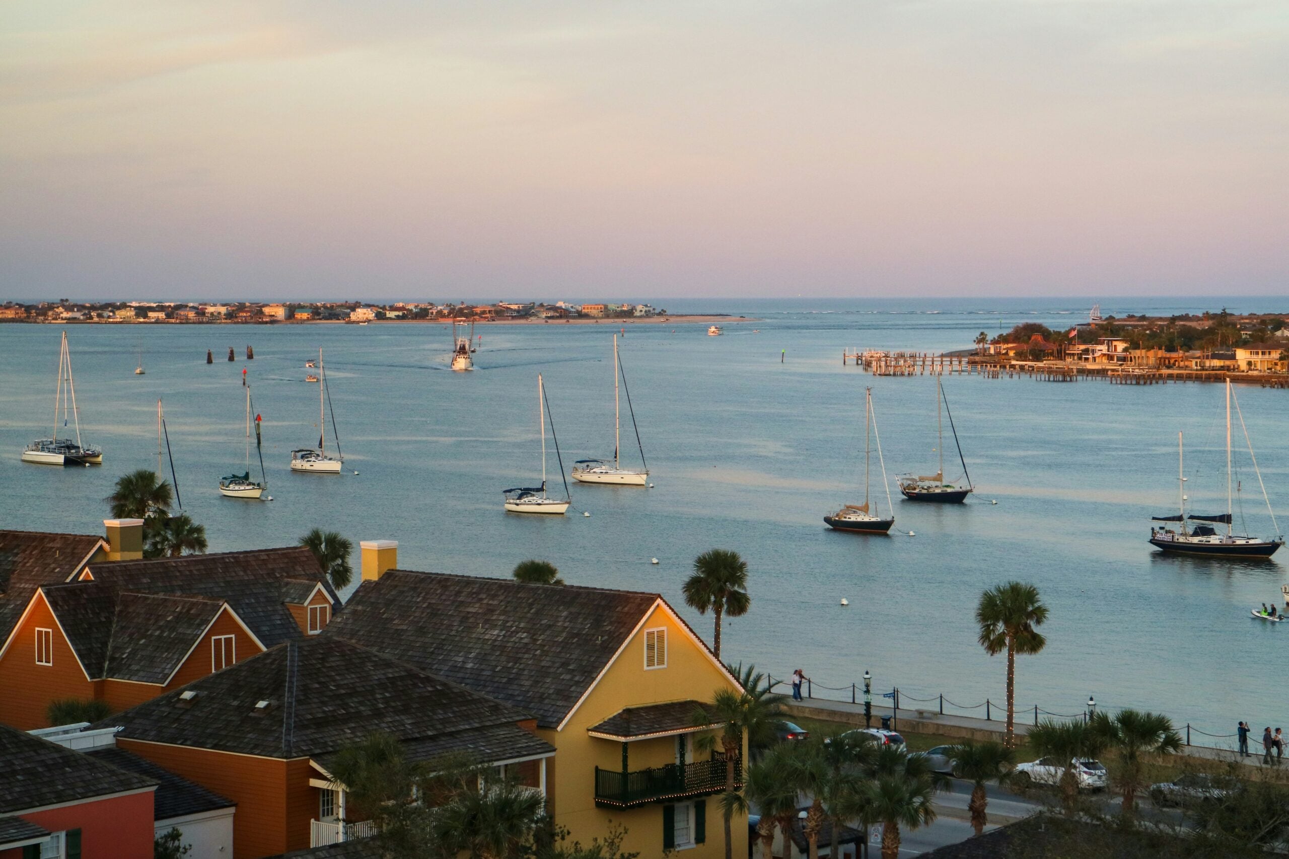 View of a calm waterfront at sunset with several sailboats anchored in the water, colorful houses in the foreground, palm trees, and a distant shoreline under a pastel sky.