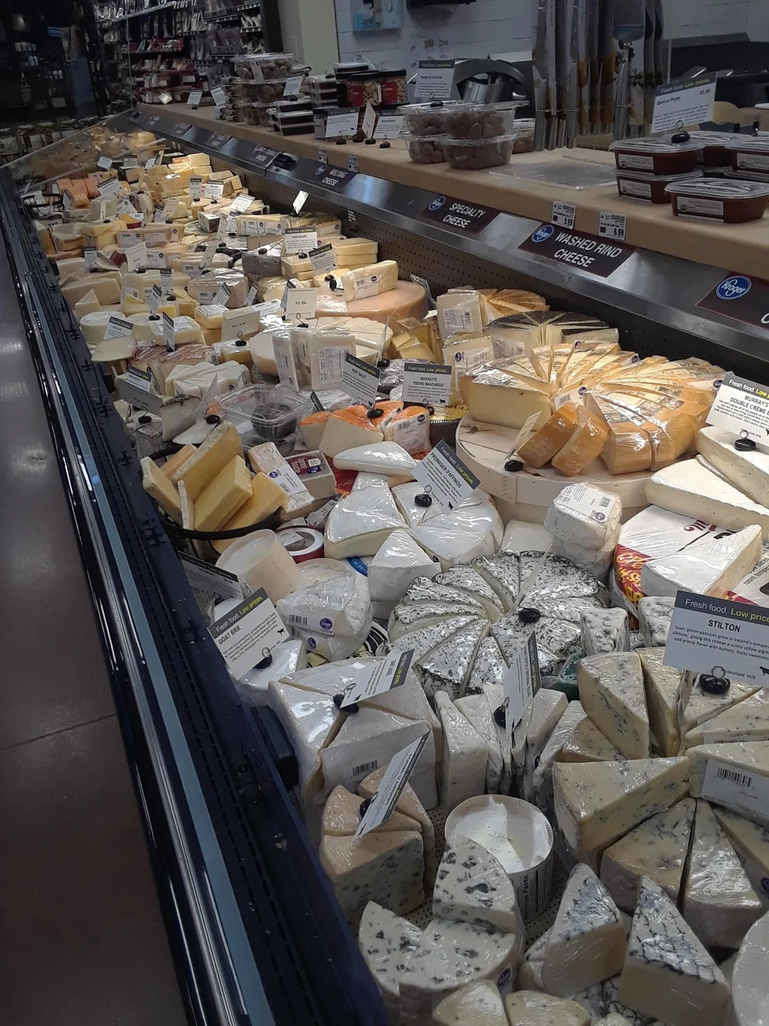 A grocery store cheese display case filled with a wide variety of cheeses, including wedges, rounds, and blocks, many with labels, stacked closely together on refrigerated shelves.