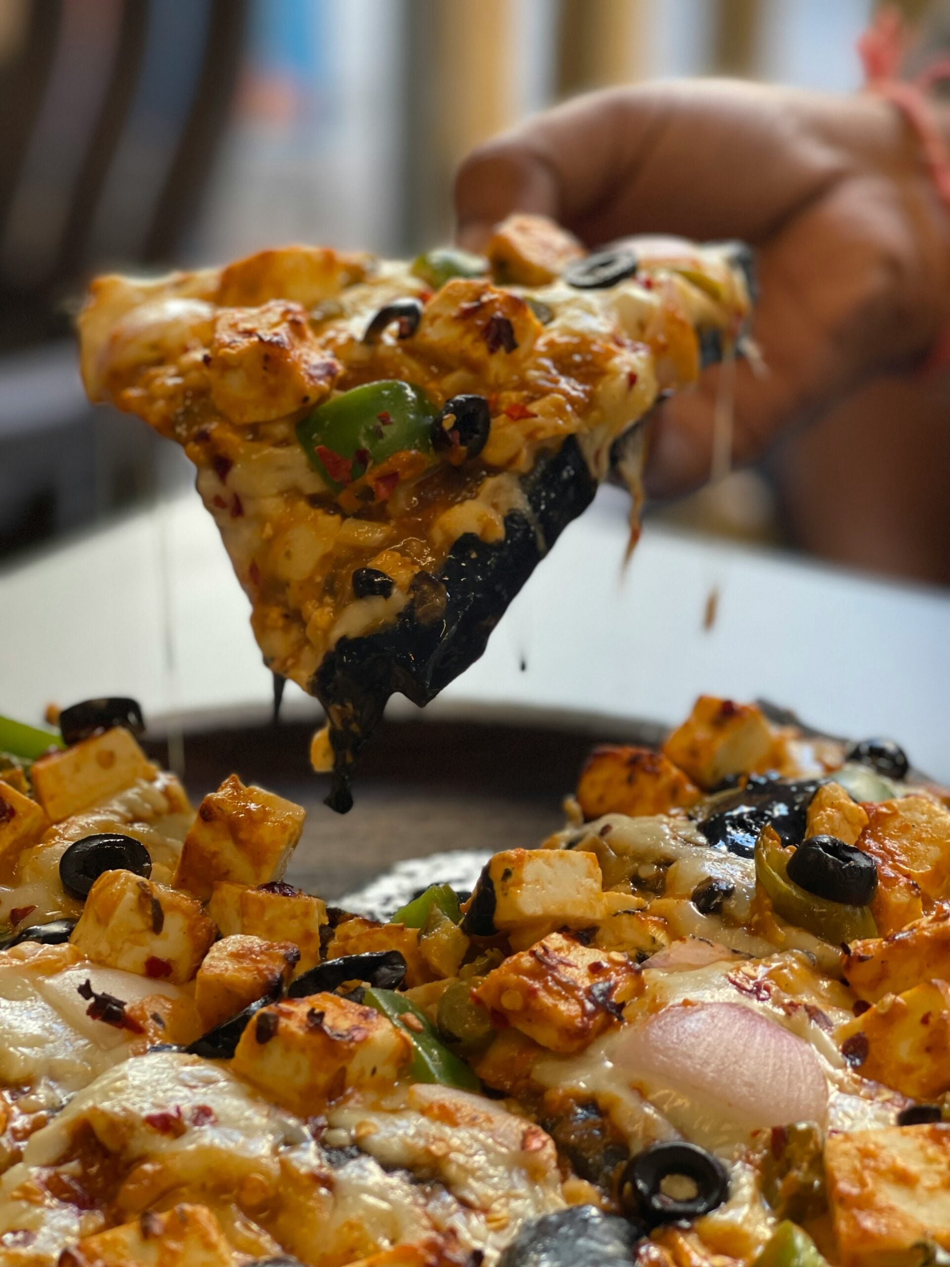 A hand lifts a cheesy slice of pizza topped with paneer, green bell peppers, onions, black olives, and red chili flakes from a whole pizza.