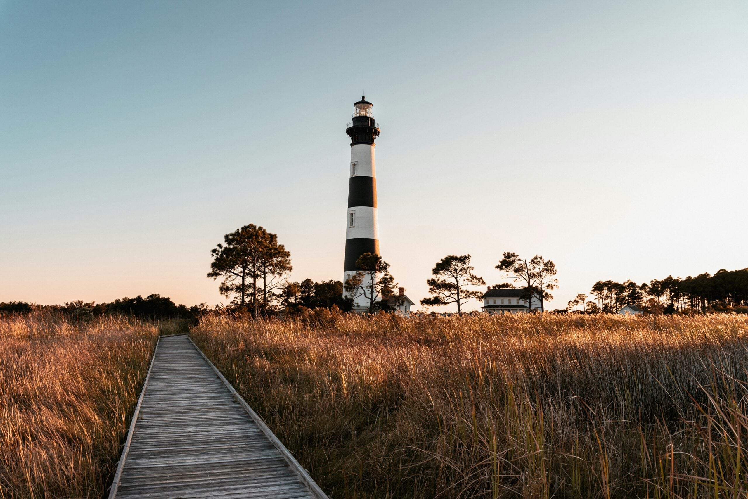 A wooden boardwalk leads through tall grass toward a black and white striped lighthouse, surrounded by trees and a small white building, under a clear sky at sunset.