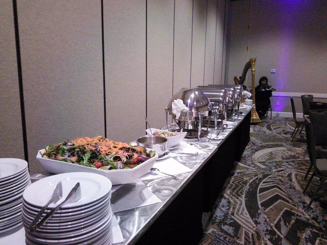 A buffet table with plates, salad, covered chafing dishes, and serving utensils is set up in a banquet room—an elegant preview of the upscale dining experiences expected from top Hotel Brands 2026. A person sits by a harp; empty chairs are visible.