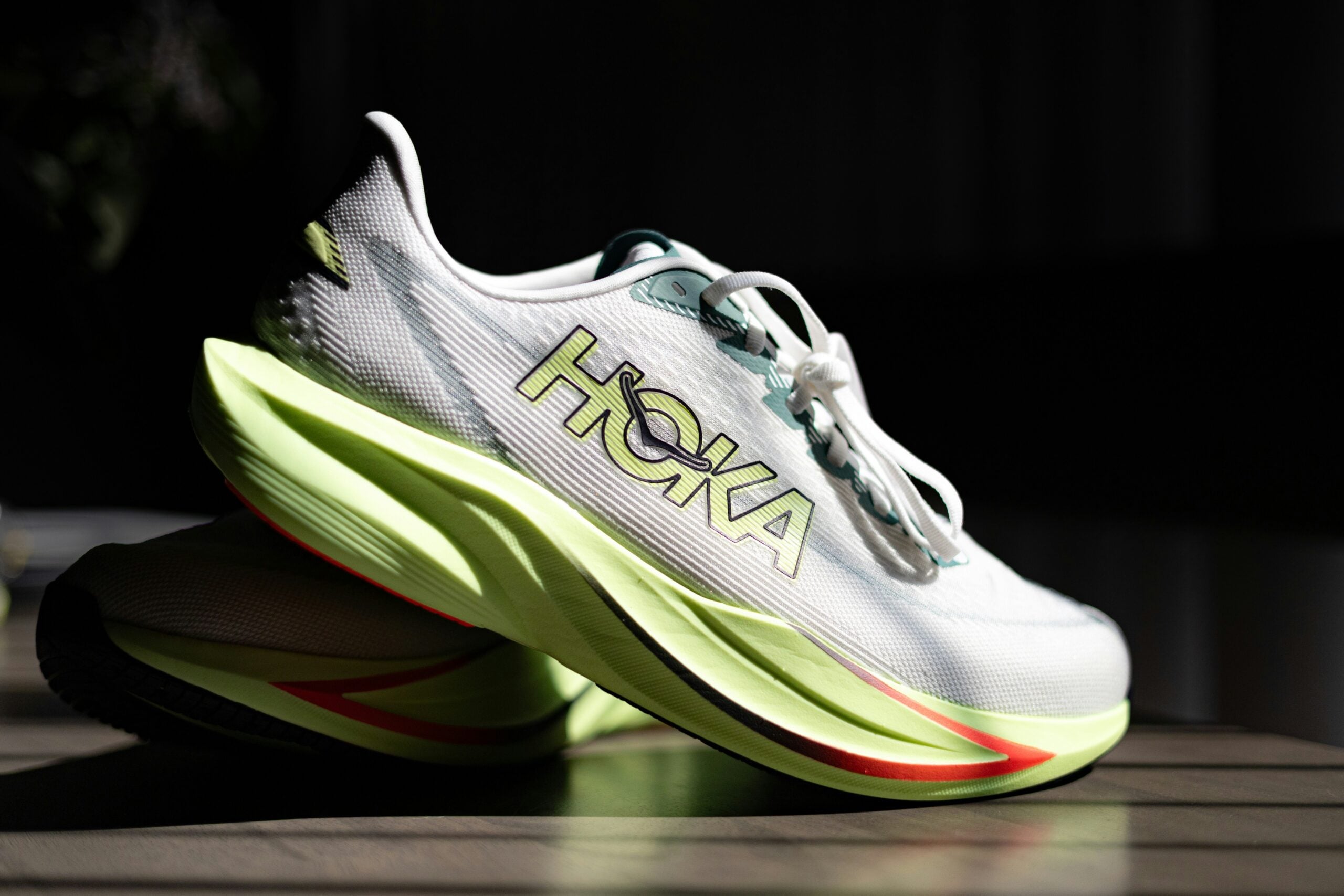 A close-up of a pair of white HOKA running shoes with neon green soles, light blue laces, and the HOKA logo on the side, placed on a wooden surface in sunlight.