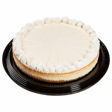 A whole plain cheesecake with a smooth white top and piped whipped cream border, placed on a round black plastic tray. The cake is pre-sliced, showing faint lines for cutting.