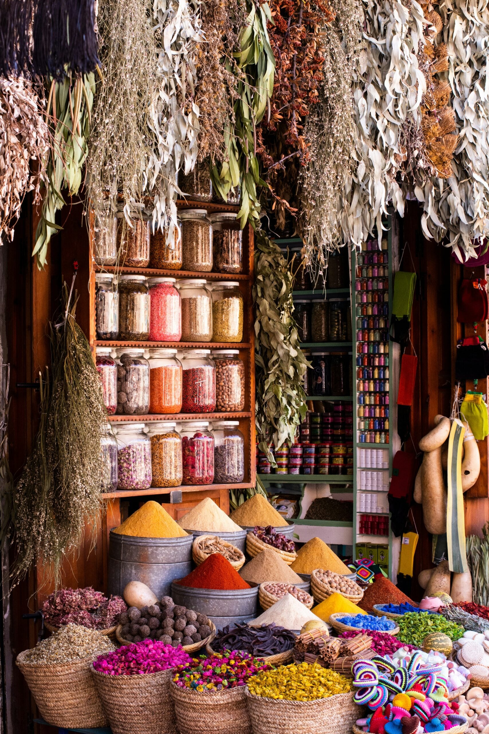Colorful spices, herbs, and dried plants in baskets and metal containers are displayed in a market stall; shelves behind hold glass jars of various preserved goods, with dried herbs hanging overhead.