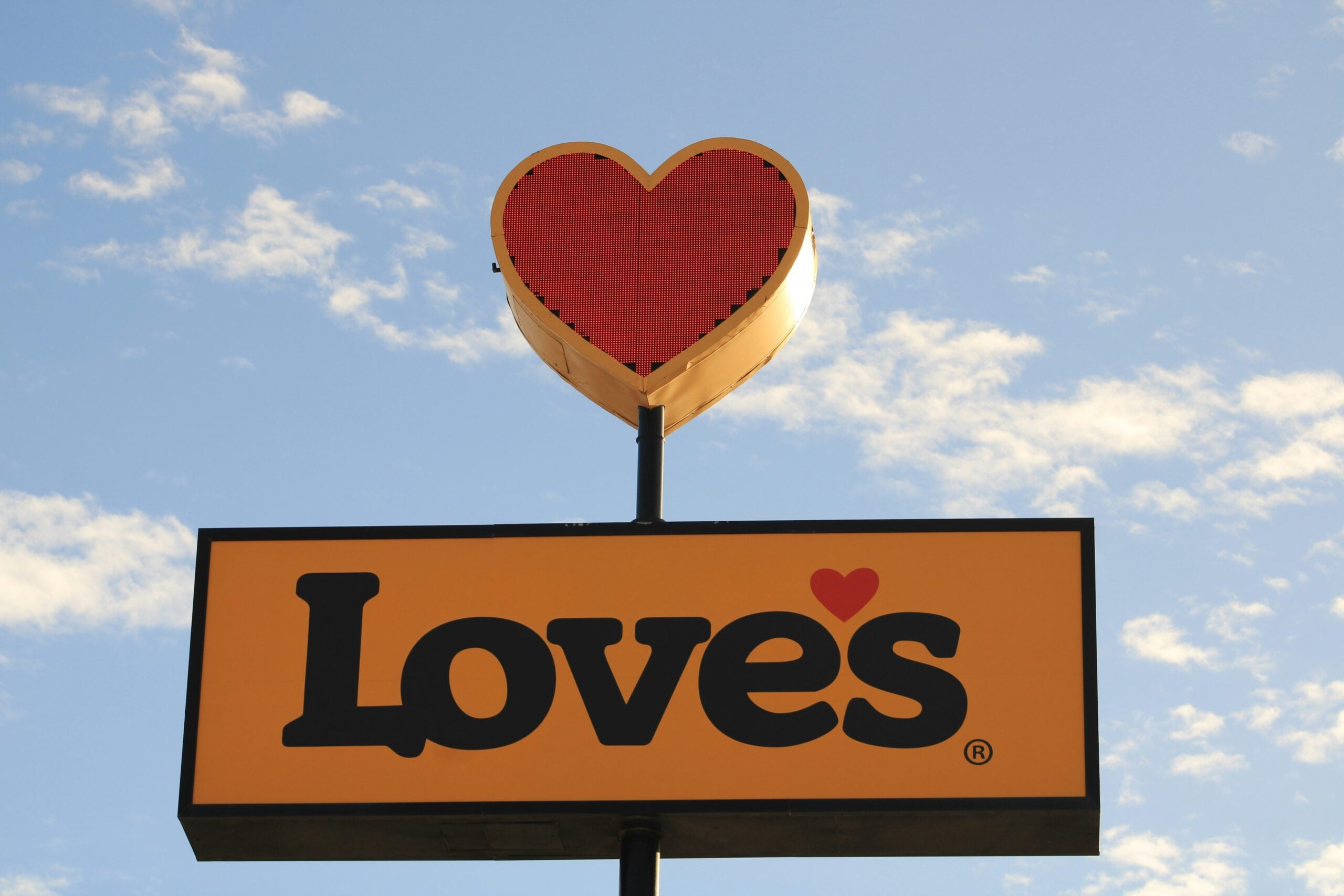A yellow sign with the word "Love’s" in black letters and a red heart above it, set against a blue sky with scattered clouds.