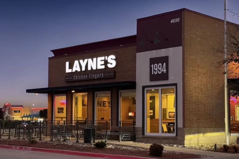 A brick restaurant with a sign reading "Layne's Chicken Fingers" and a large "1994" on the front. The building is lit up at dusk with outdoor seating and some cars visible in the background.