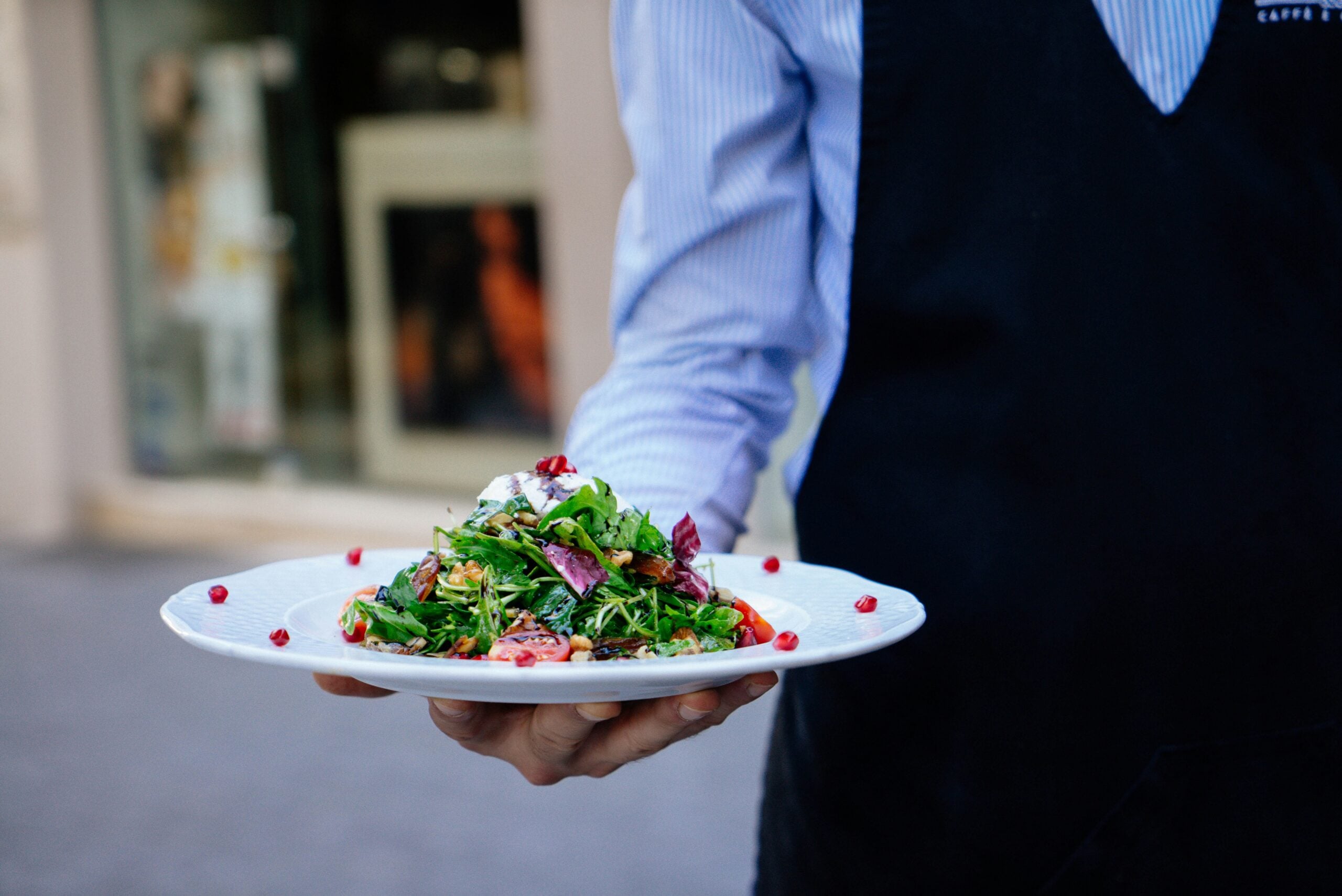 A person in a blue shirt and black apron holds a white plate with a colorful salad topped with greens, pomegranate seeds, and cheese, outdoors near a building.