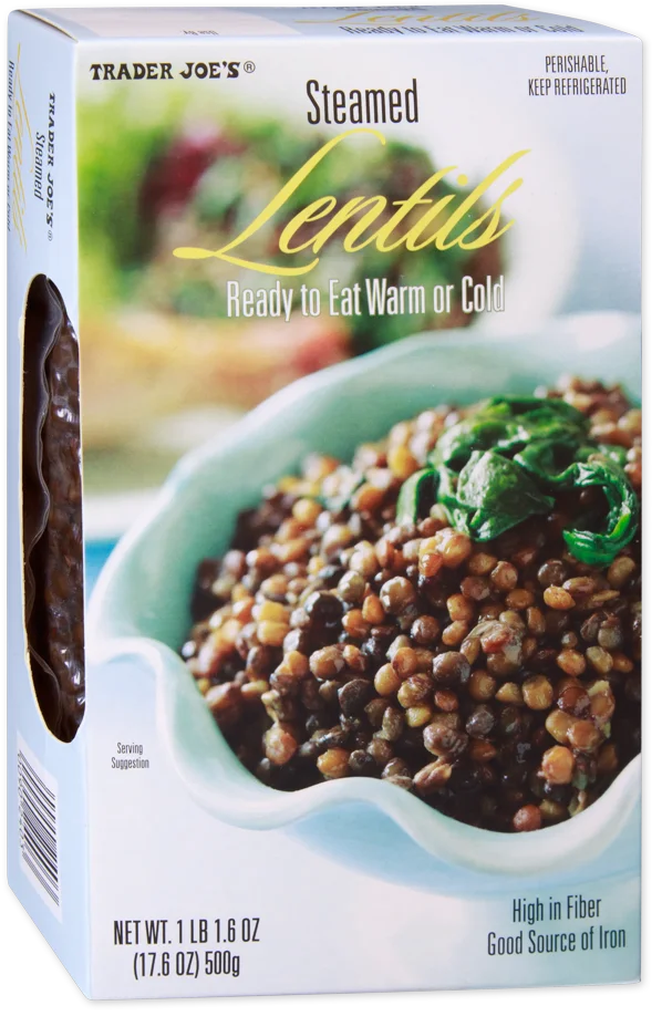 A box of Trader Joe’s Steamed Lentils shows a bowl of cooked lentils garnished with greens. Text highlights they are ready to eat warm or cold, high in fiber, a good source of iron, and the net weight is 500g.
