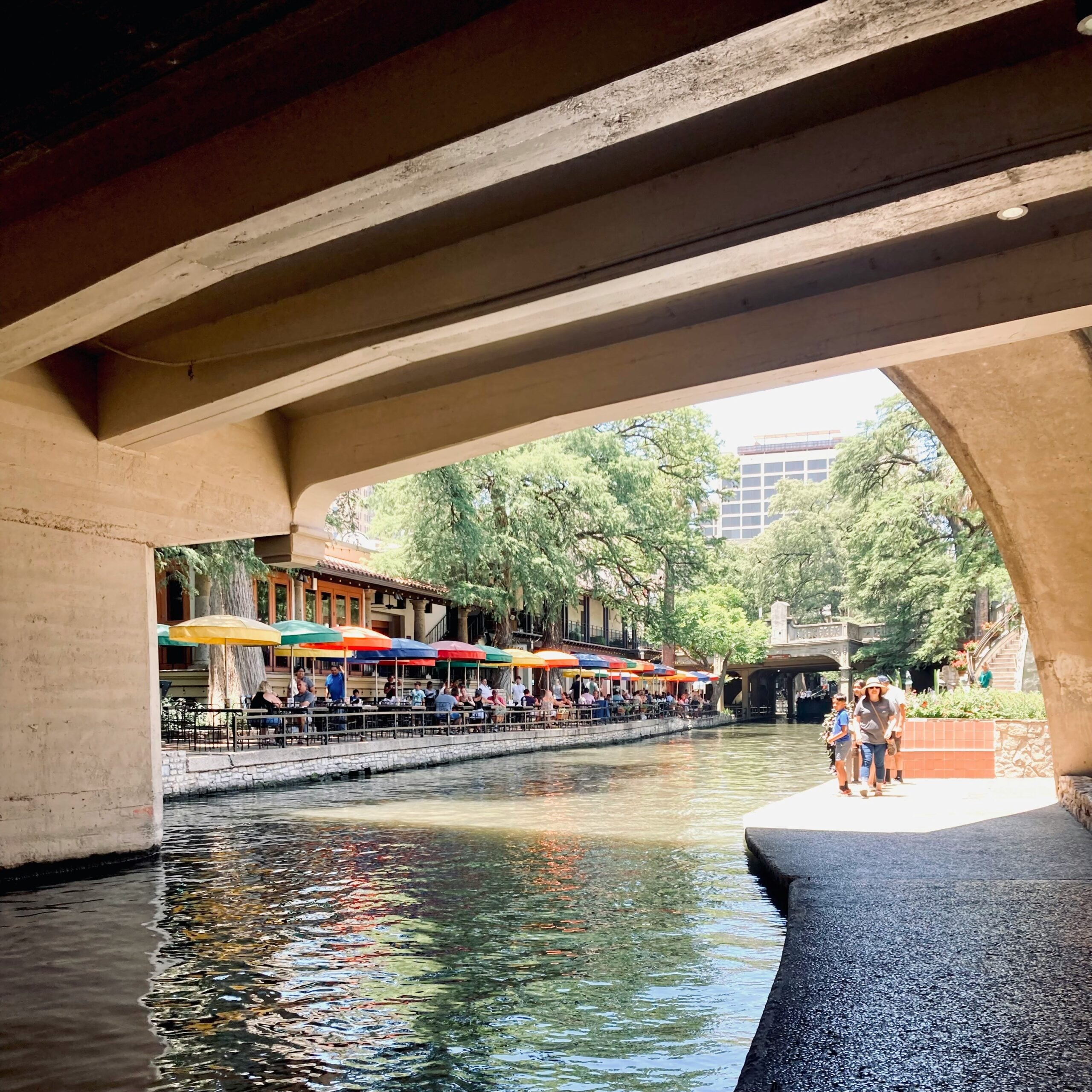 A riverwalk scene under a bridge with people walking along the water, colorful umbrellas shading outdoor diners, trees lining the walkway, and buildings in the background on a sunny day.