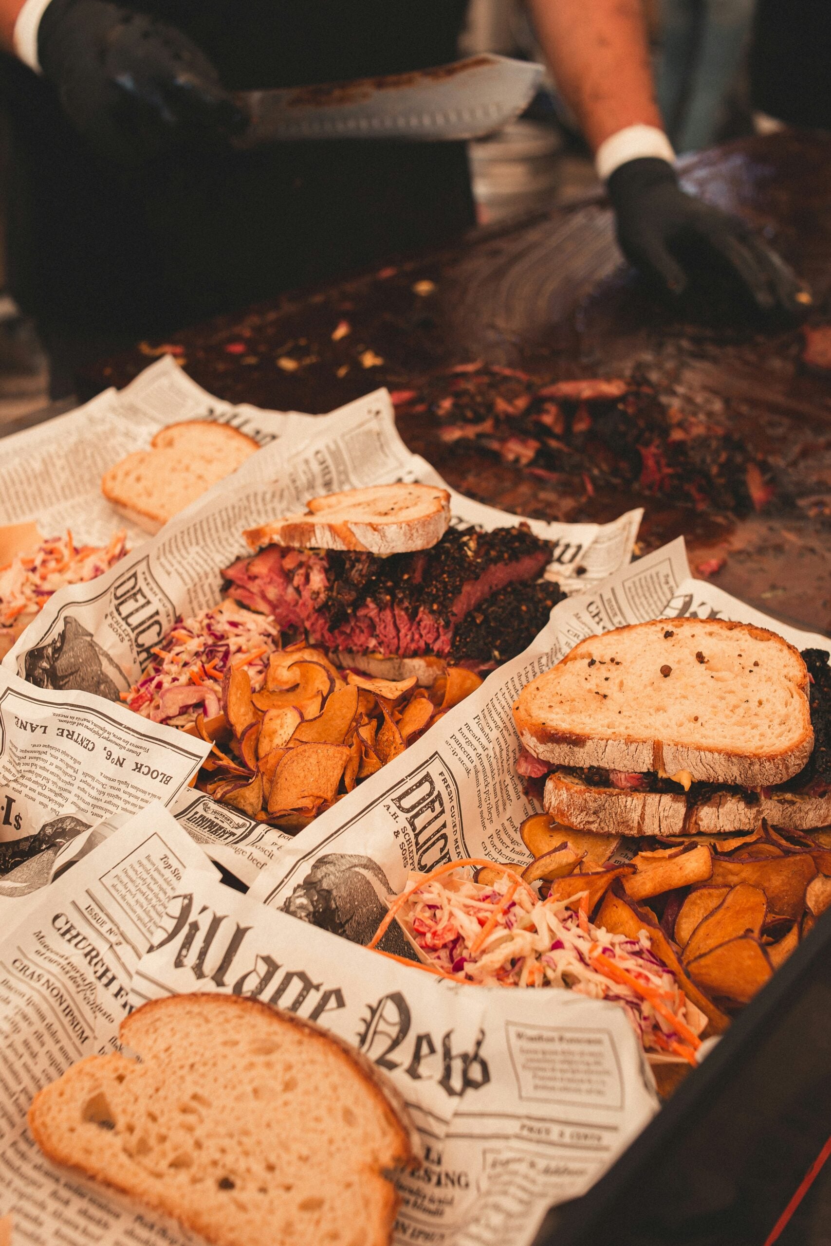 Two trays lined with newspaper print paper hold sandwiches with thick slices of meat, coleslaw, and chips. A person wearing black gloves is slicing more meat at a wooden counter in the background.