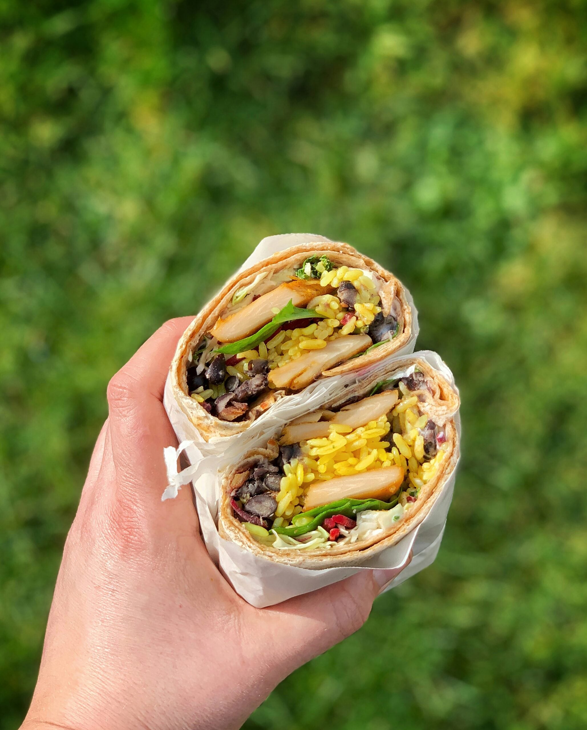 A hand holds a burrito cut in half, showing layers of rice, beans, vegetables, and possibly tofu or grilled chicken, with a blurred green background.
