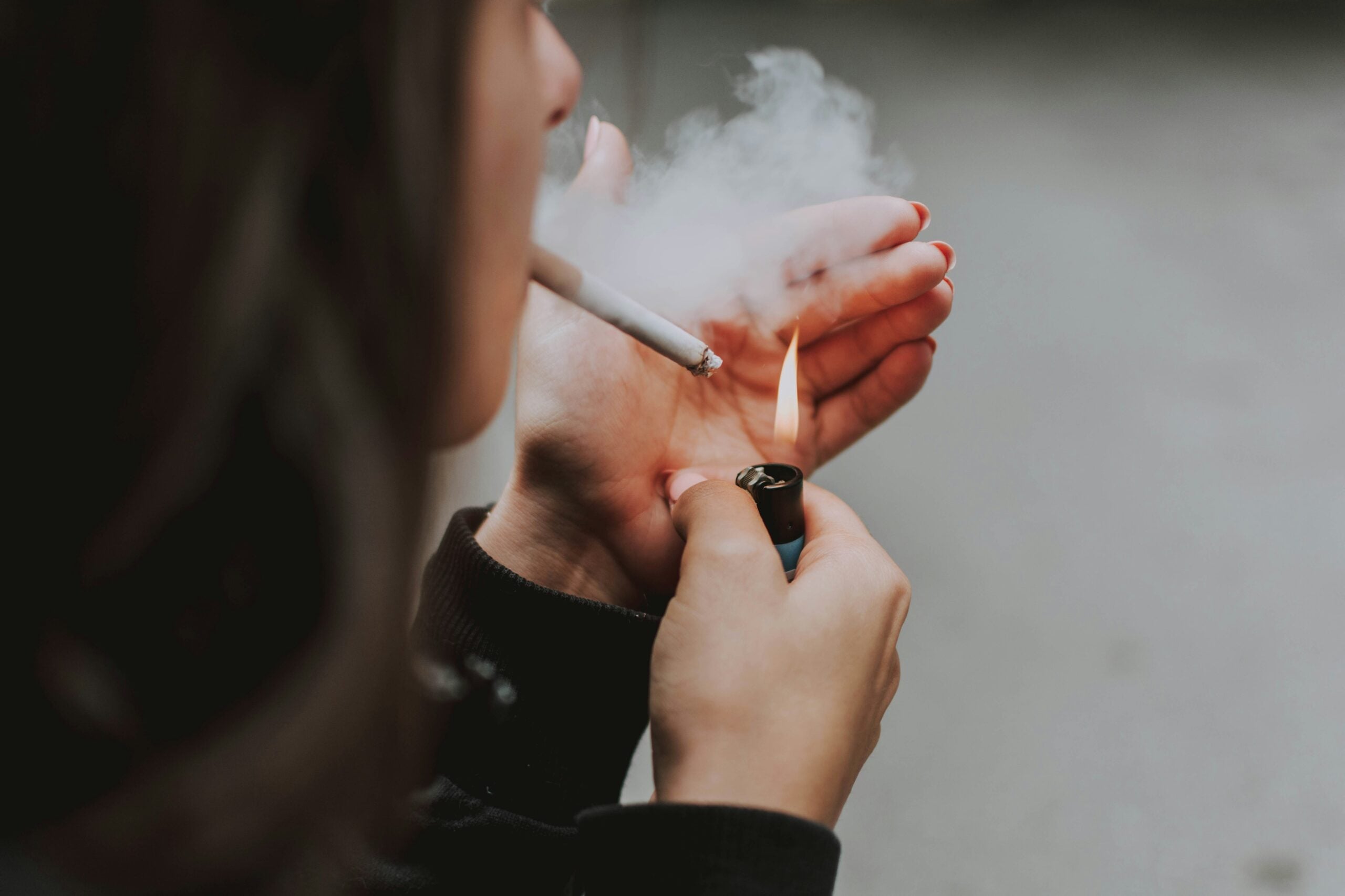 A person holds a cigarette in their mouth and uses a lighter to ignite it, shielding the flame with their hand. Smoke is visible near the cigarette. The background is blurred.
