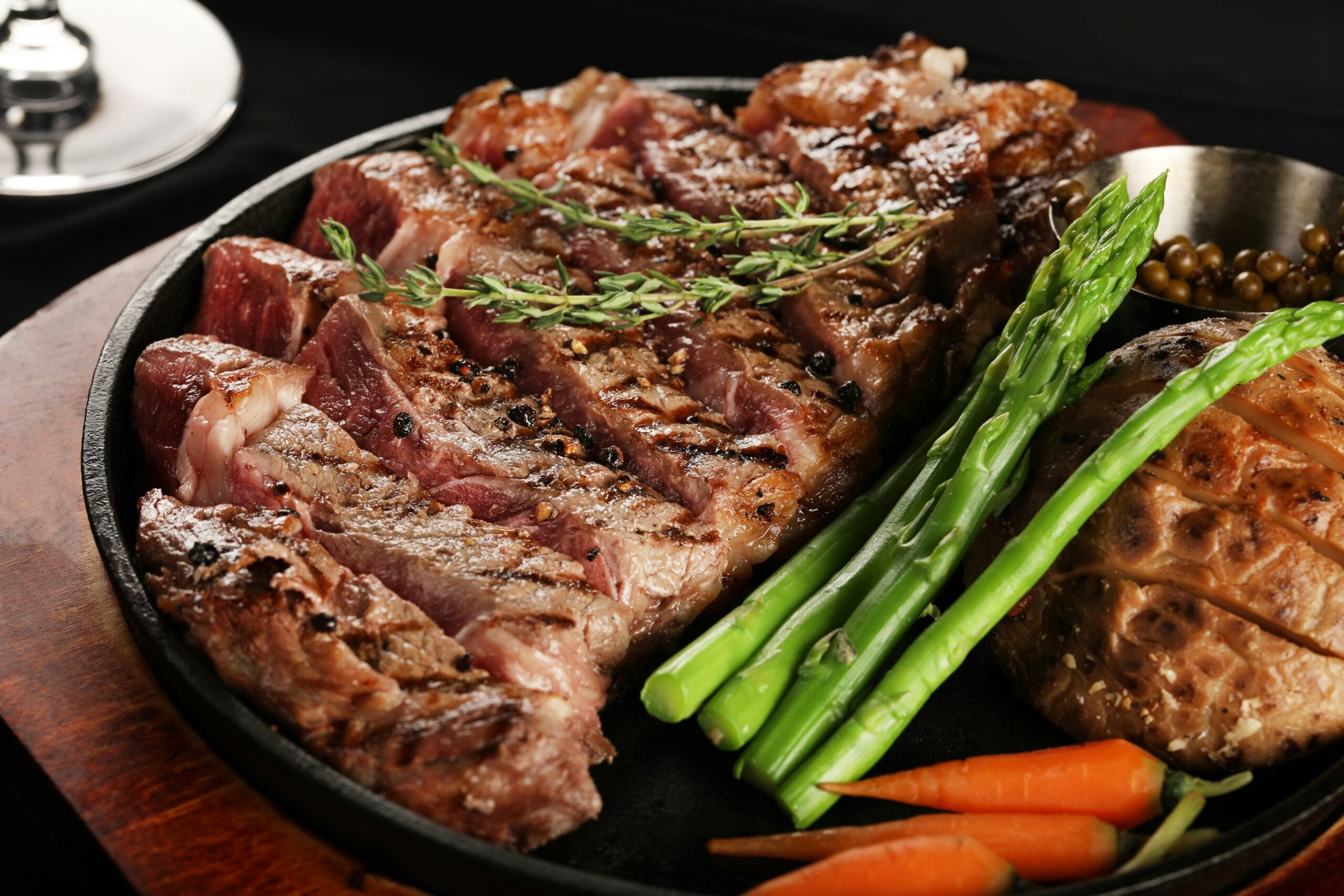 A grilled steak sliced into strips, garnished with fresh rosemary, served with asparagus spears, whole roasted garlic, green peppercorns, and small carrots on a dark plate.