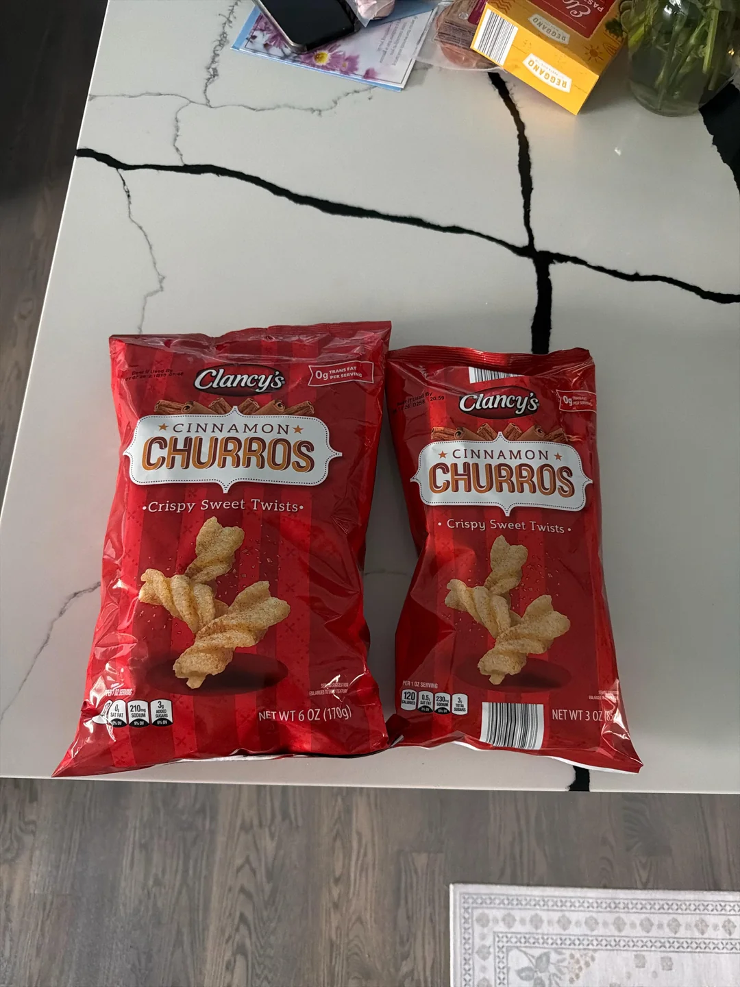 Two red bags of Clancy’s Cinnamon Churros, labeled "Crispy Sweet Twists," sit on a white marble table with dark cracks. Other groceries are partially visible in the background.