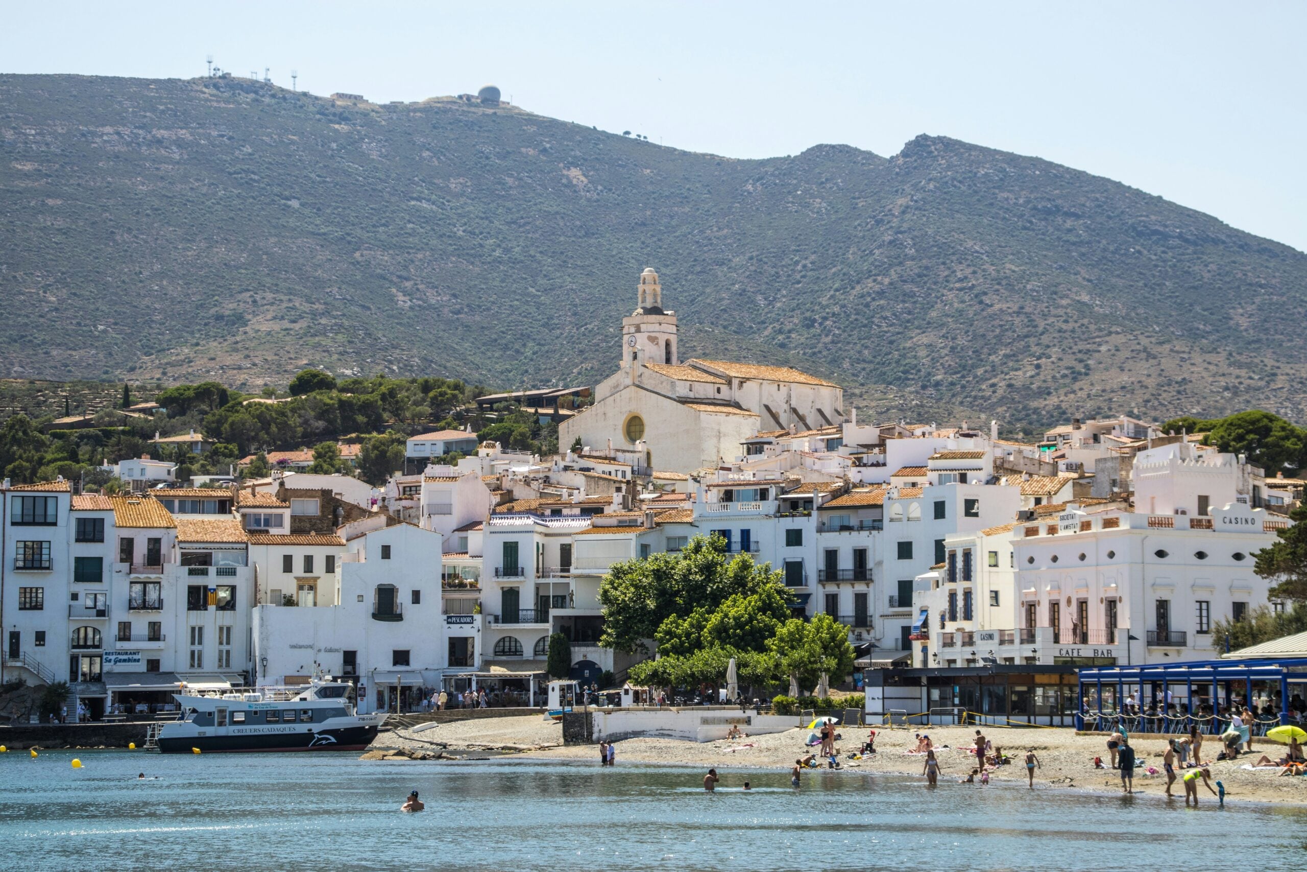 A coastal town with white buildings and a church on a hill, set against a backdrop of green mountains. People are swimming and relaxing on the beach, and boats are docked near the shore.