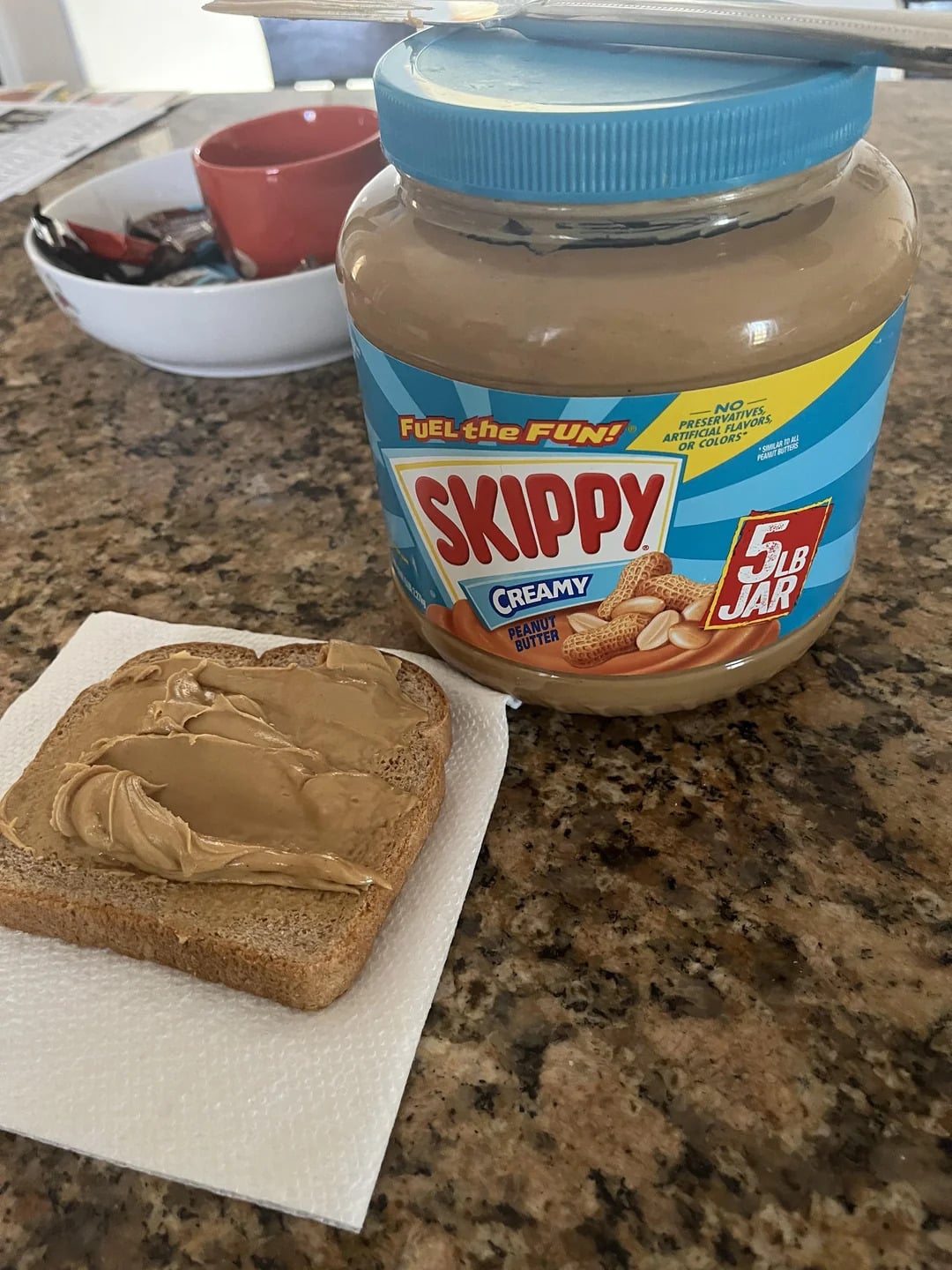 A large jar of Skippy creamy peanut butter, one of the best peanut butter brands, sits on a granite counter next to a slice of bread with peanut butter spread on it, resting on a napkin. In the background, there is a bowl with various items.