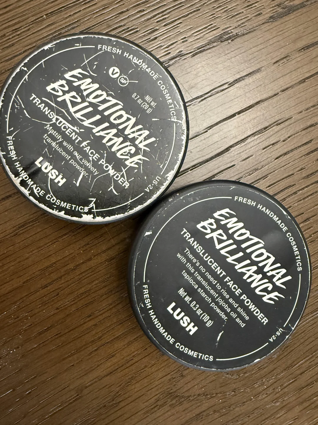 Two round black containers of Lush Emotional Brilliance translucent face powder sit on a wooden surface. The white text on the lids displays the product's name and details.
