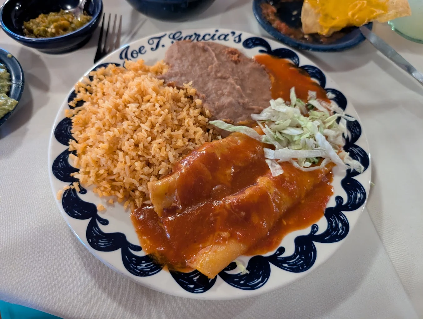 A plate of Mexican food with two cheese enchiladas covered in red sauce, refried beans, Mexican rice, and shredded lettuce on a decorative plate. Other small dishes are partially visible around the plate.