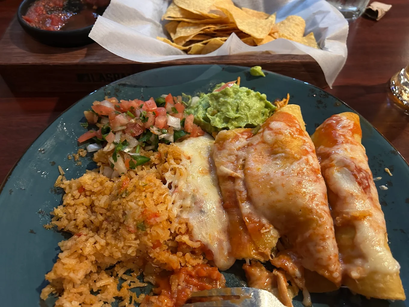 A plate with two cheese enchiladas, Mexican rice, pico de gallo, and guacamole. In the background, there is a basket of tortilla chips and a bowl of red salsa.