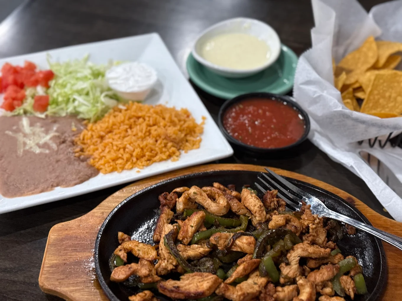 A sizzling plate of chicken fajitas with green peppers, served with rice, refried beans, lettuce, tomatoes, sour cream, and sides of salsa, queso dip, and tortilla chips on a dark table.