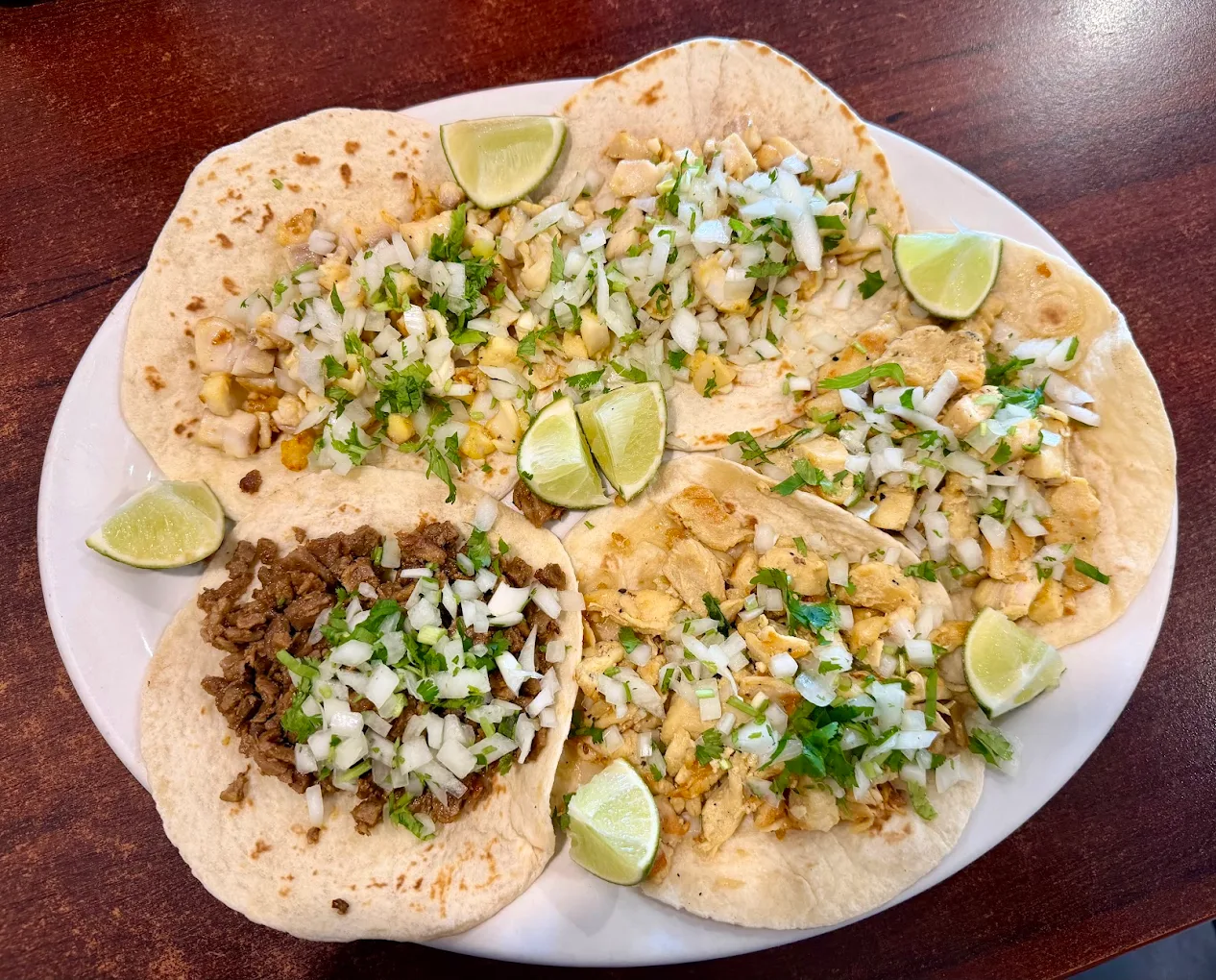 A plate with five tacos topped with chopped onions and cilantro; four tacos appear to have grilled chicken, while one has beef. Lime wedges are placed on the plate. The food is on a wooden table.