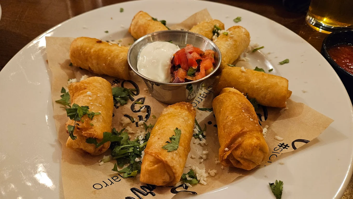 Seven golden-brown taquitos arranged in a circle on a plate, garnished with chopped cilantro and cheese, served with small containers of sour cream and pico de gallo in the center.