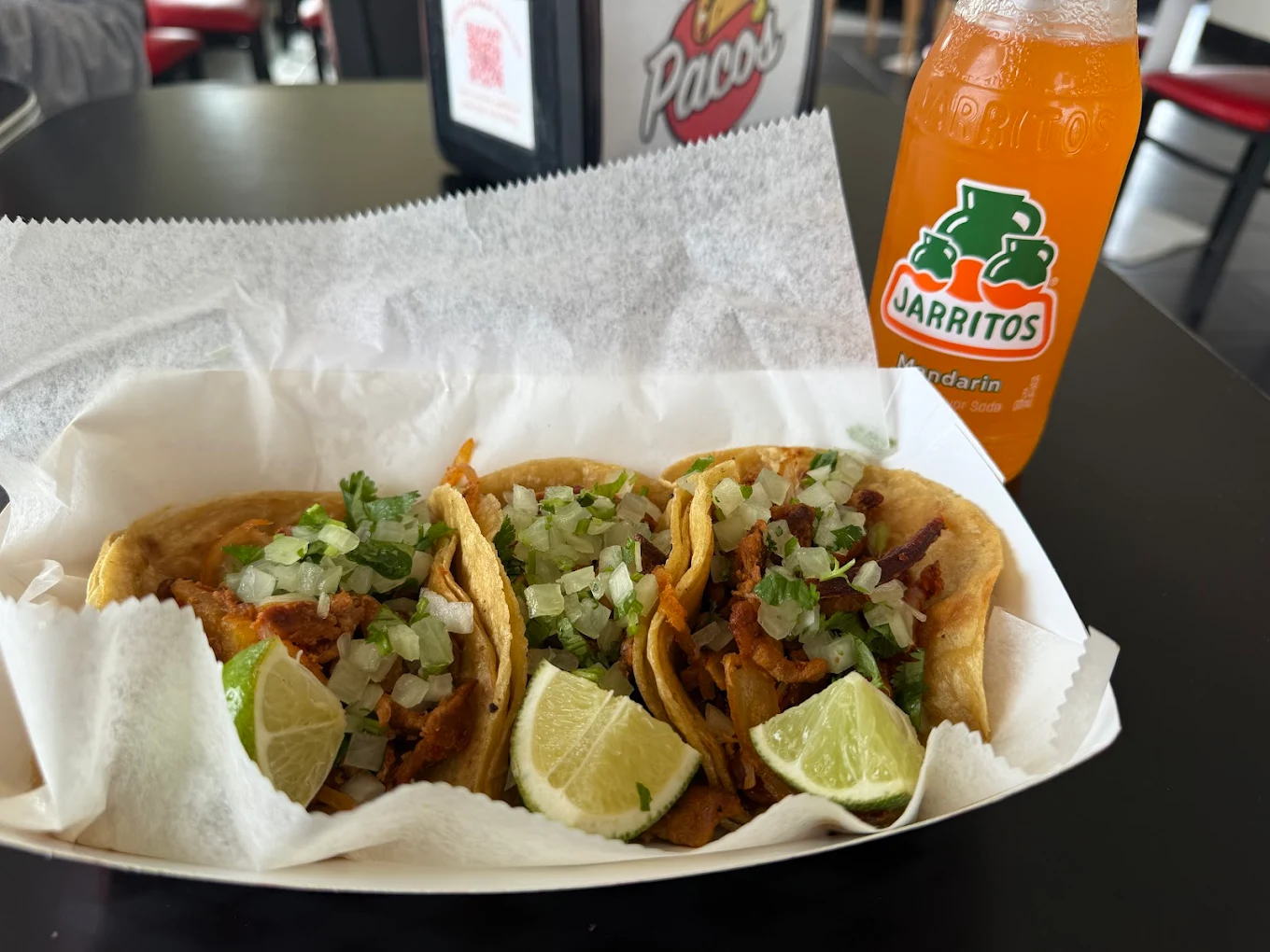 Three tacos filled with meat, chopped onions, and cilantro are served in a paper tray with lime wedges. An orange Jarritos soda bottle is next to the tray on a black table in a casual restaurant setting.