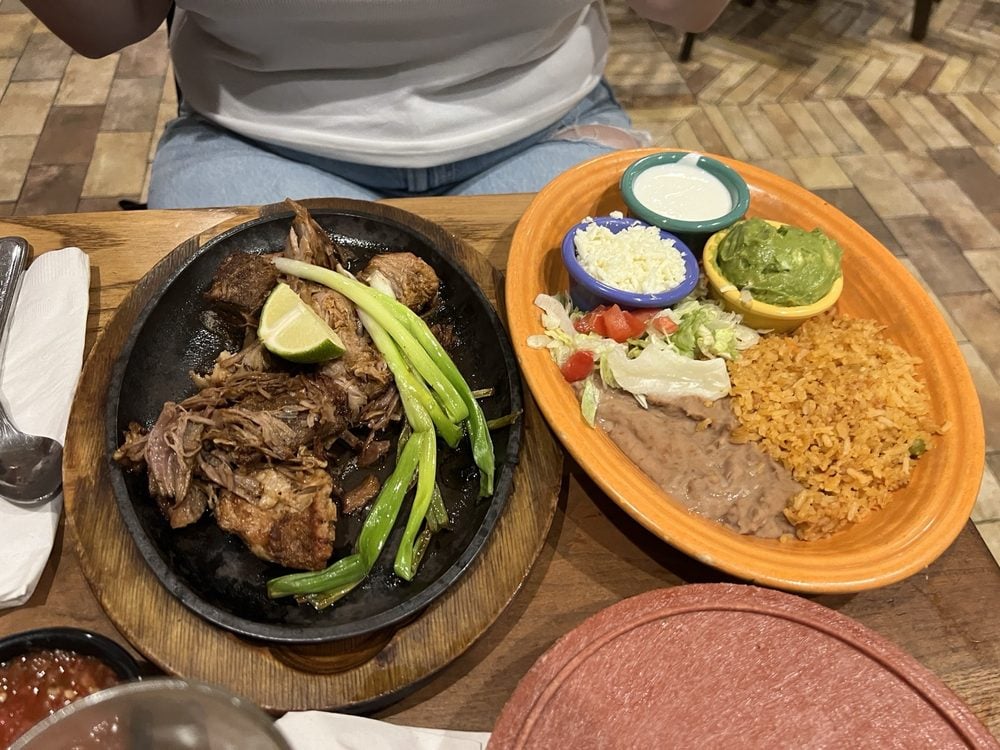 A plate of shredded beef with grilled green onions and a lime wedge is next to a platter of Mexican rice, refried beans, guacamole, sour cream, cheese, lettuce, and tomato. A person sits behind the food.