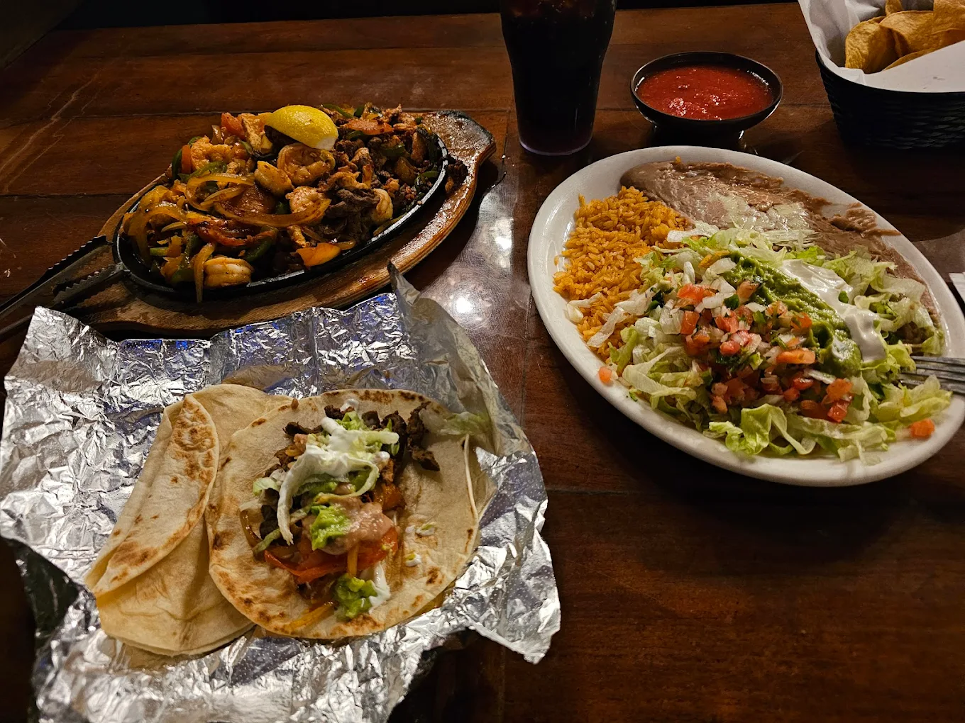 A table with sizzling fajitas, a plate of rice, refried beans, salad topped with sour cream, salsa, foil-wrapped tacos filled with meat, and a drink. A basket of tortilla chips is in the background.