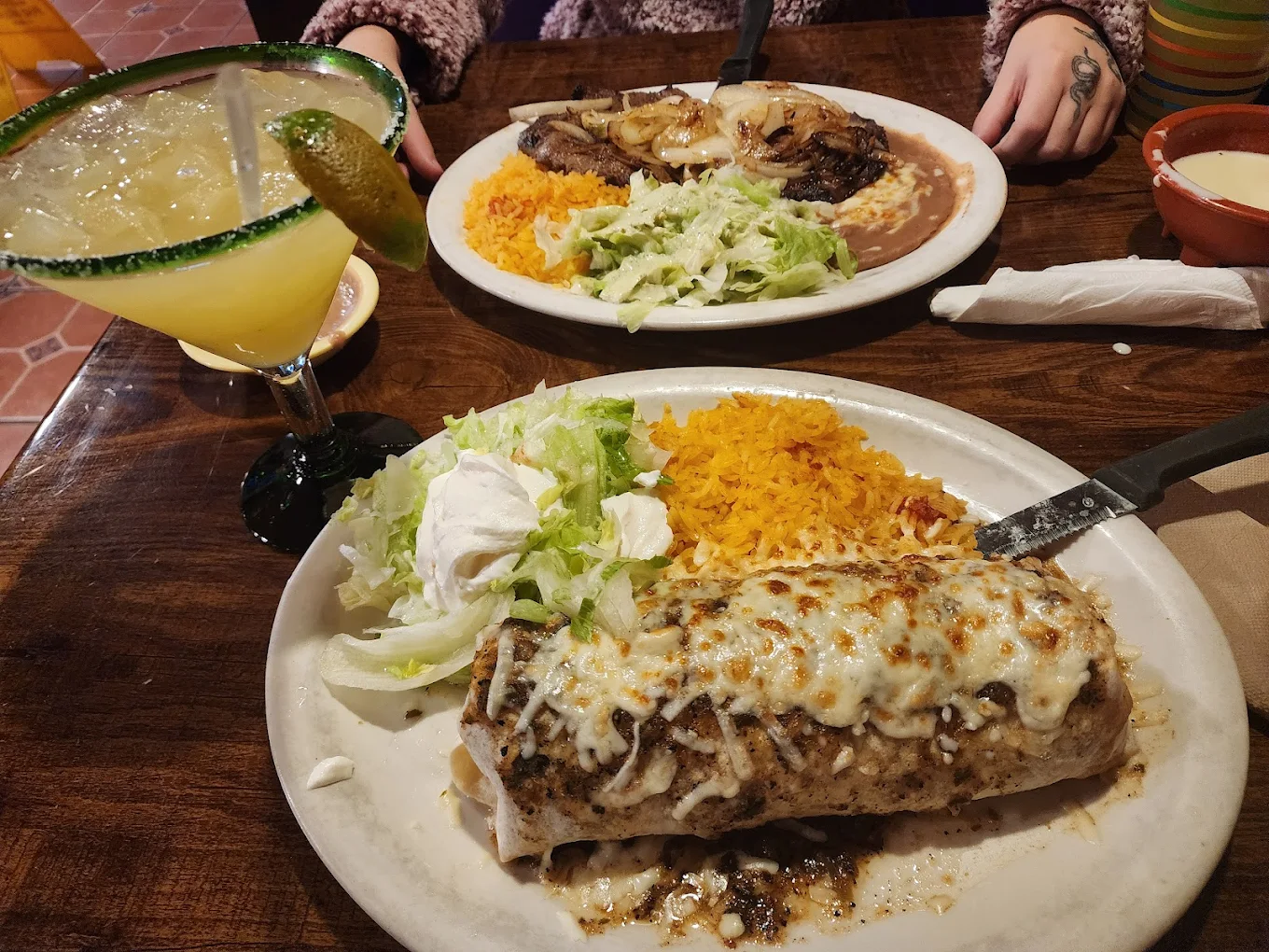 A plate with a cheesy burrito, lettuce, sour cream, and Mexican rice sits on a wooden table next to a margarita. Another plate with grilled meat, onions, beans, rice, and salad is across the table.