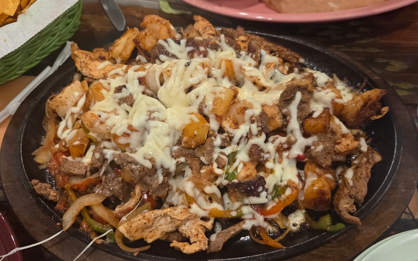 A sizzling platter of fajitas with grilled chicken, beef, and vegetables, topped with melted cheese, served on a black skillet.
