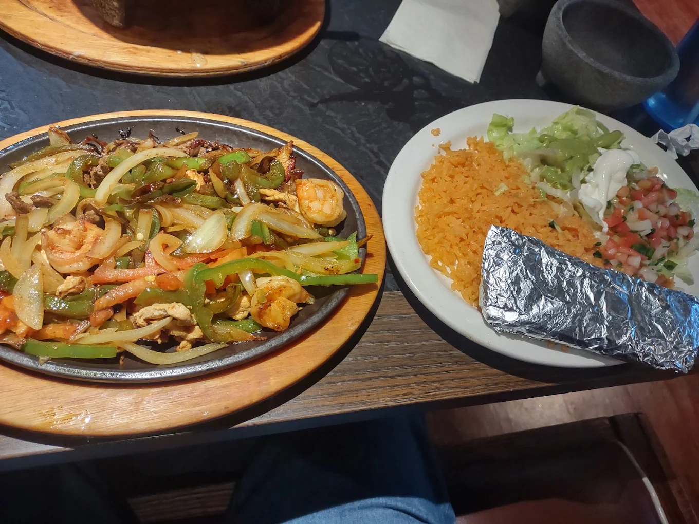 A sizzling platter of fajitas with shrimp, chicken, onions, and bell peppers sits next to a plate with orange rice, lettuce, sour cream, pico de gallo, and a foil-wrapped tortilla on a dark table.