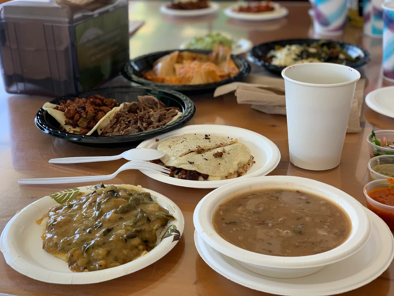 A table with plates of Mexican food, including tacos, gorditas, a dish with melted cheese and peppers, a bowl of beans, a white cup, plastic utensils, and various salsas.