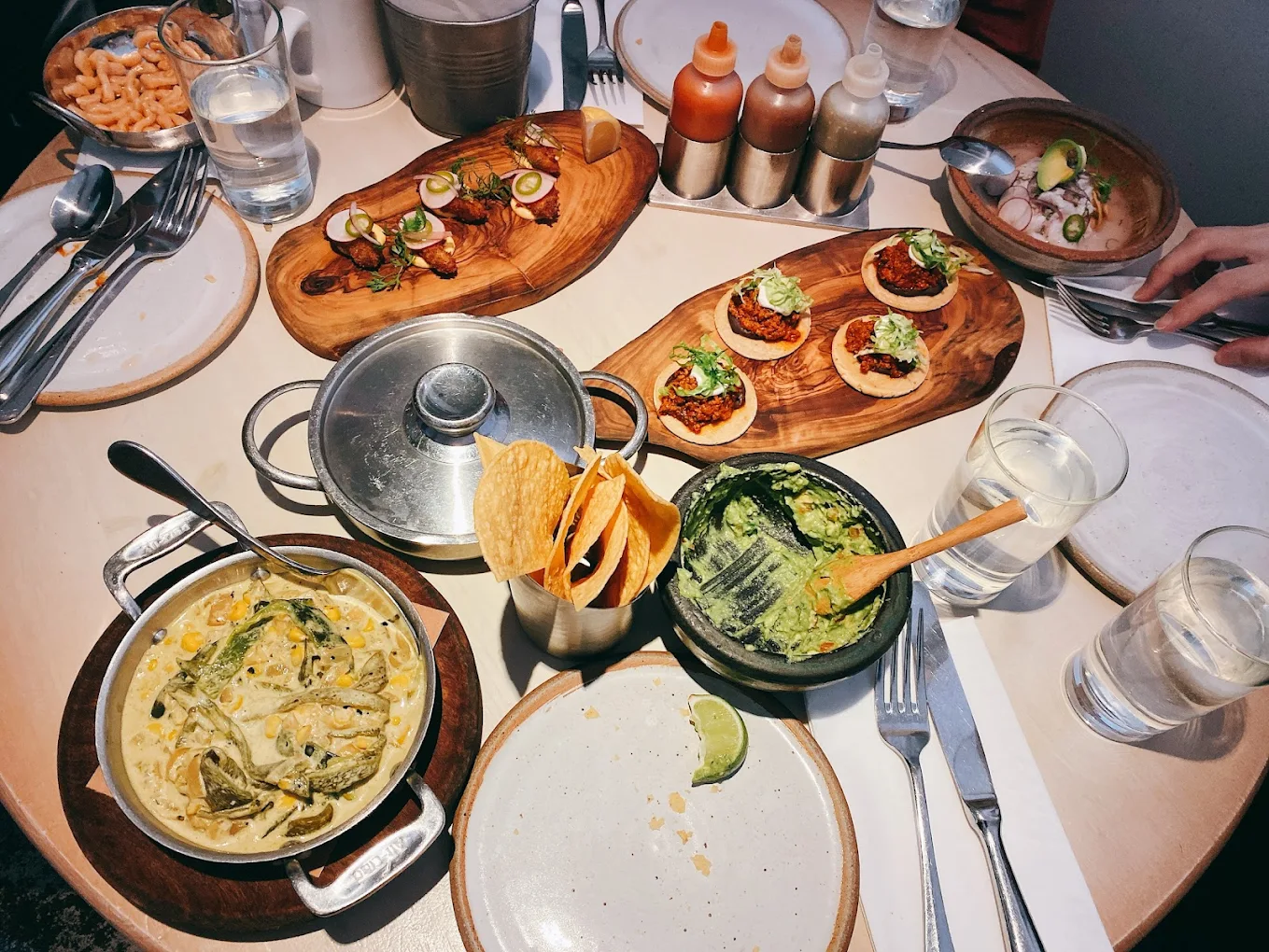 A table set with various Mexican dishes, including tacos, guacamole with tortilla chips, and other small plates. There are also sauces, drinks, and empty plates with utensils around the table.