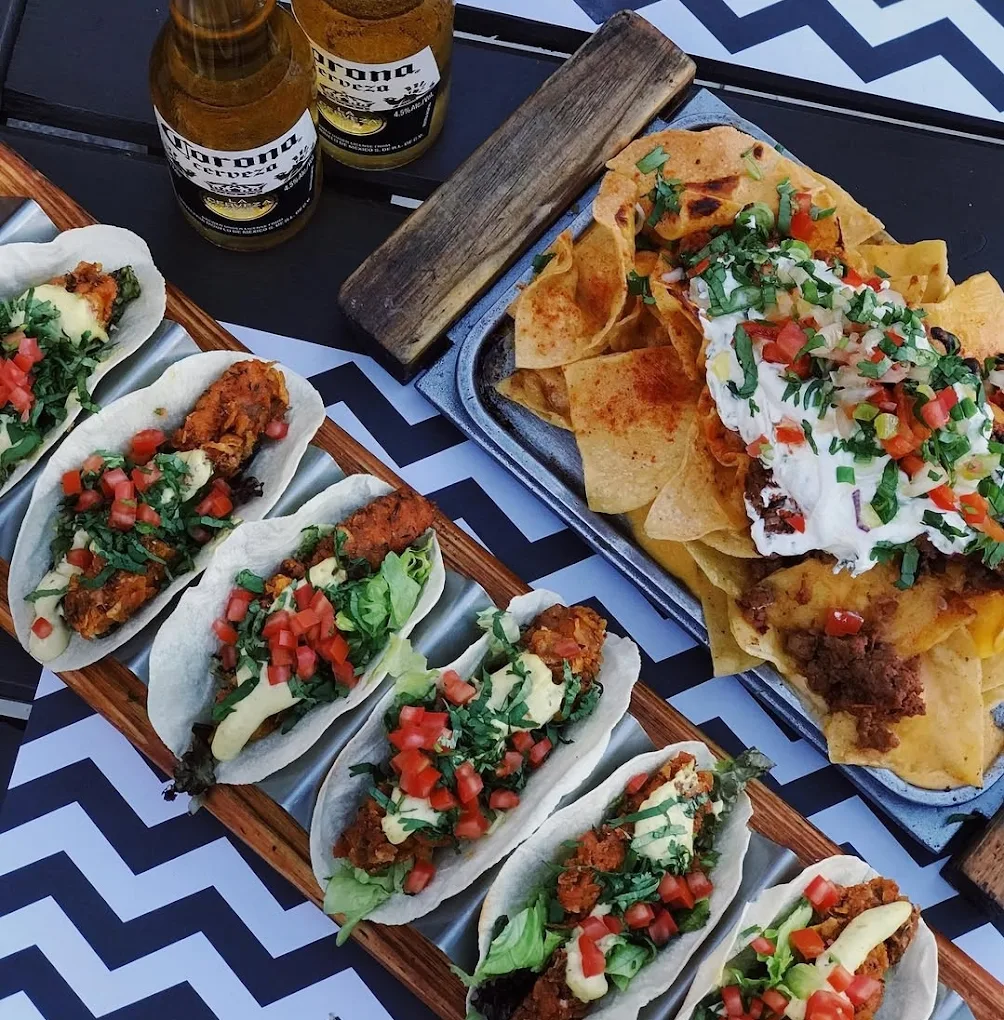 A tray of chicken tacos topped with lettuce, avocado, tomato, and cilantro is next to a plate of nachos with cheese, sour cream, and salsa. Two bottles of Corona beer are nearby on a zigzag-patterned table.