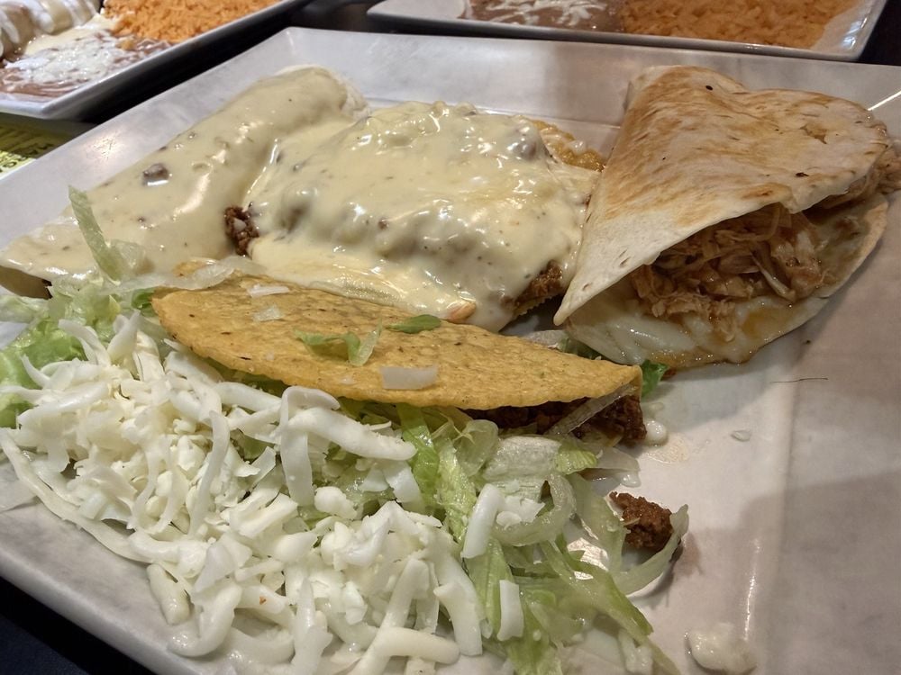 A plate with a hard shell taco, a soft taco, an enchilada topped with white cheese sauce, and a side of shredded lettuce and cheese. Rice is visible in the background.