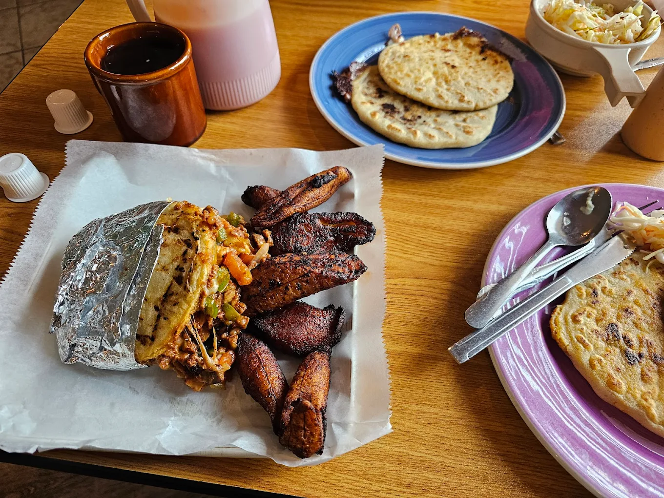 A meal with grilled plantains, a foil-wrapped sandwich filled with meat and vegetables, and corn tortillas (pupusas) on plates, with utensils, a mug, and sauce bottles on a wooden table.