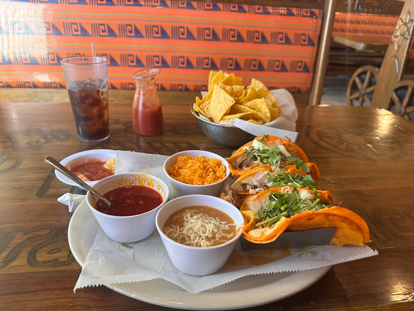 A table set with birria tacos topped with cilantro and onions, sides of rice and beans, a bowl of consommé, tortilla chips with salsa, a glass of soda, and a jar of salsa in a colorful restaurant setting.