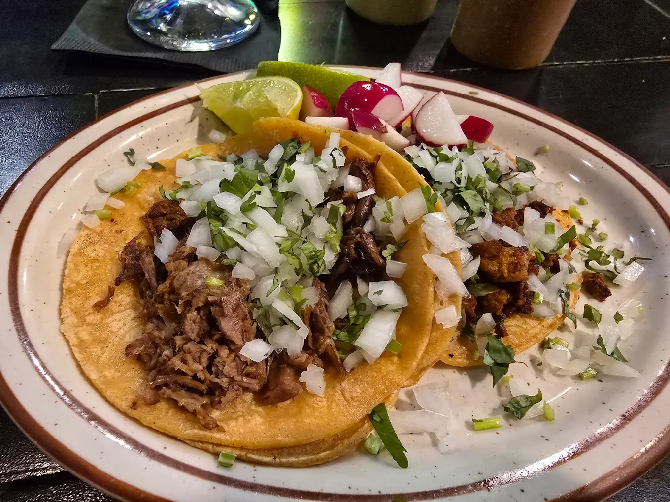 Two tacos filled with chopped meat, diced onions, and cilantro on a plate, served with lime wedges and sliced radishes on the side. The plate is set on a dark tabletop.