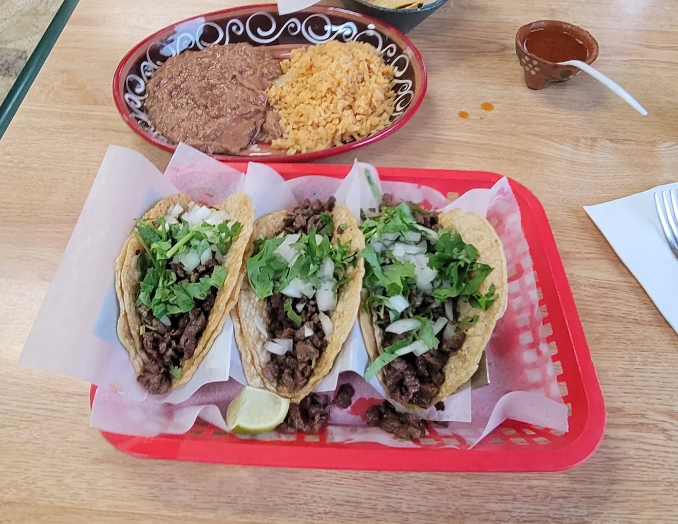 Three beef street tacos topped with chopped onions and cilantro are served on a red tray with lime wedges. In the background, a plate of refried beans and Mexican rice is visible, along with a small bowl of salsa.