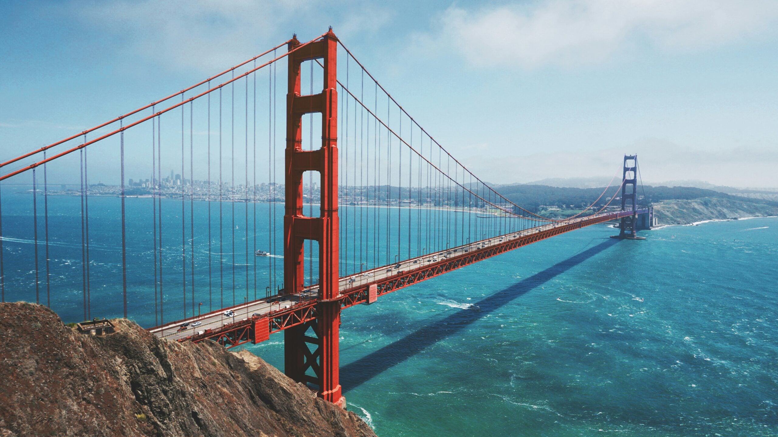 A wide view of the Golden Gate Bridge stretching over blue water, with the city of San Francisco visible in the background under a partly cloudy sky.
