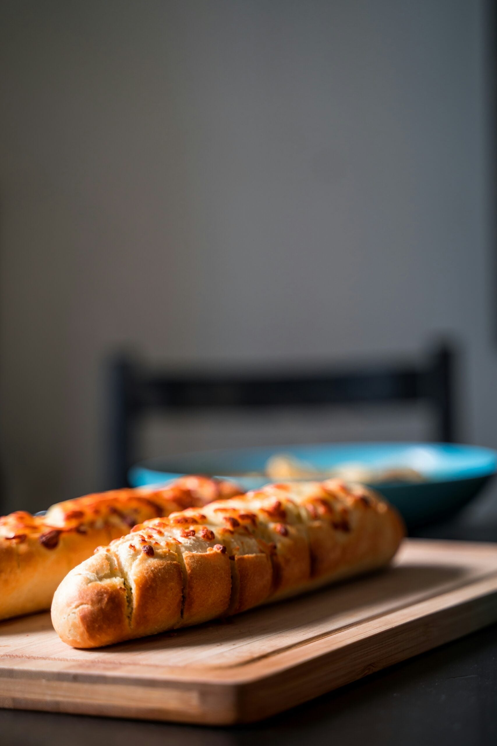 Two loaves of garlic bread with golden, crispy tops sit on a wooden cutting board, with a blurred blue dish and chair in the background. The scene is softly lit, emphasizing the bread’s texture.