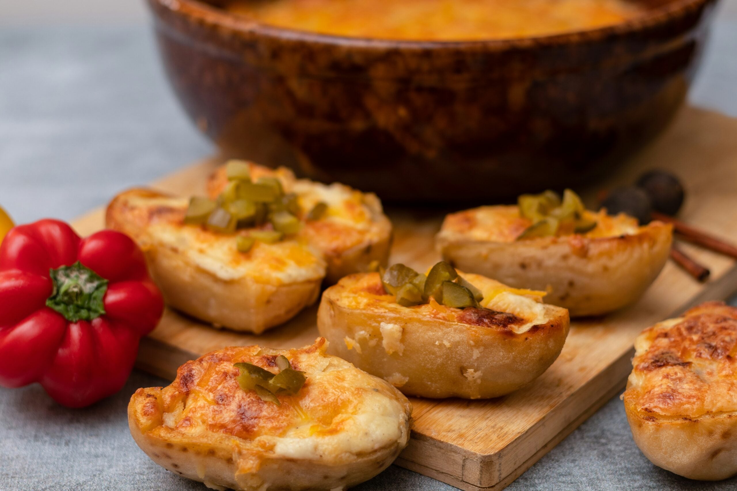 A close-up of baked stuffed potato halves topped with melted cheese and chopped jalapeños on a wooden board, with a red bell pepper and a brown baking dish in the background.
