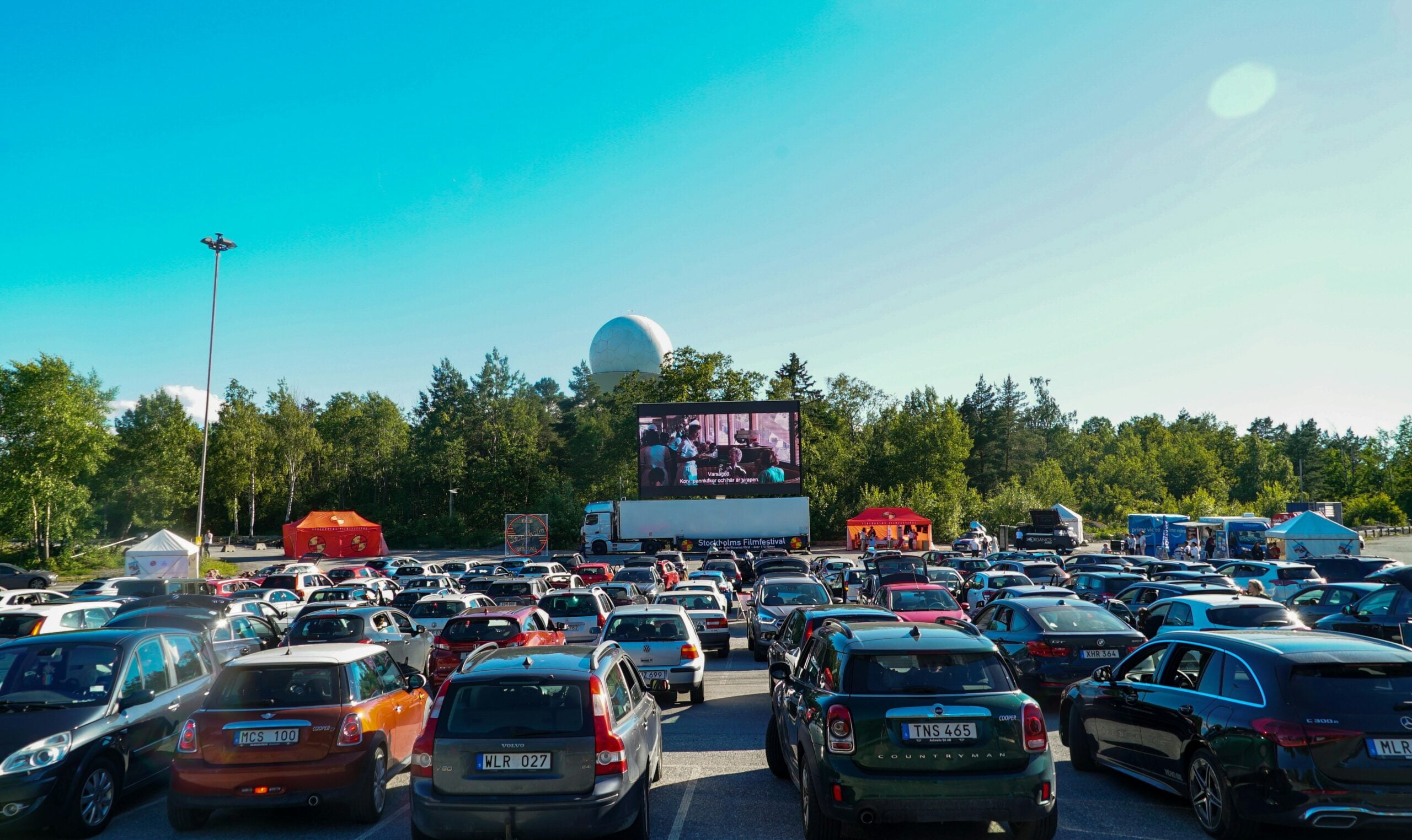 Rows of cars parked at an outdoor drive-in movie theater, all facing a large screen displaying a film, with trees and a clear blue sky in the background.