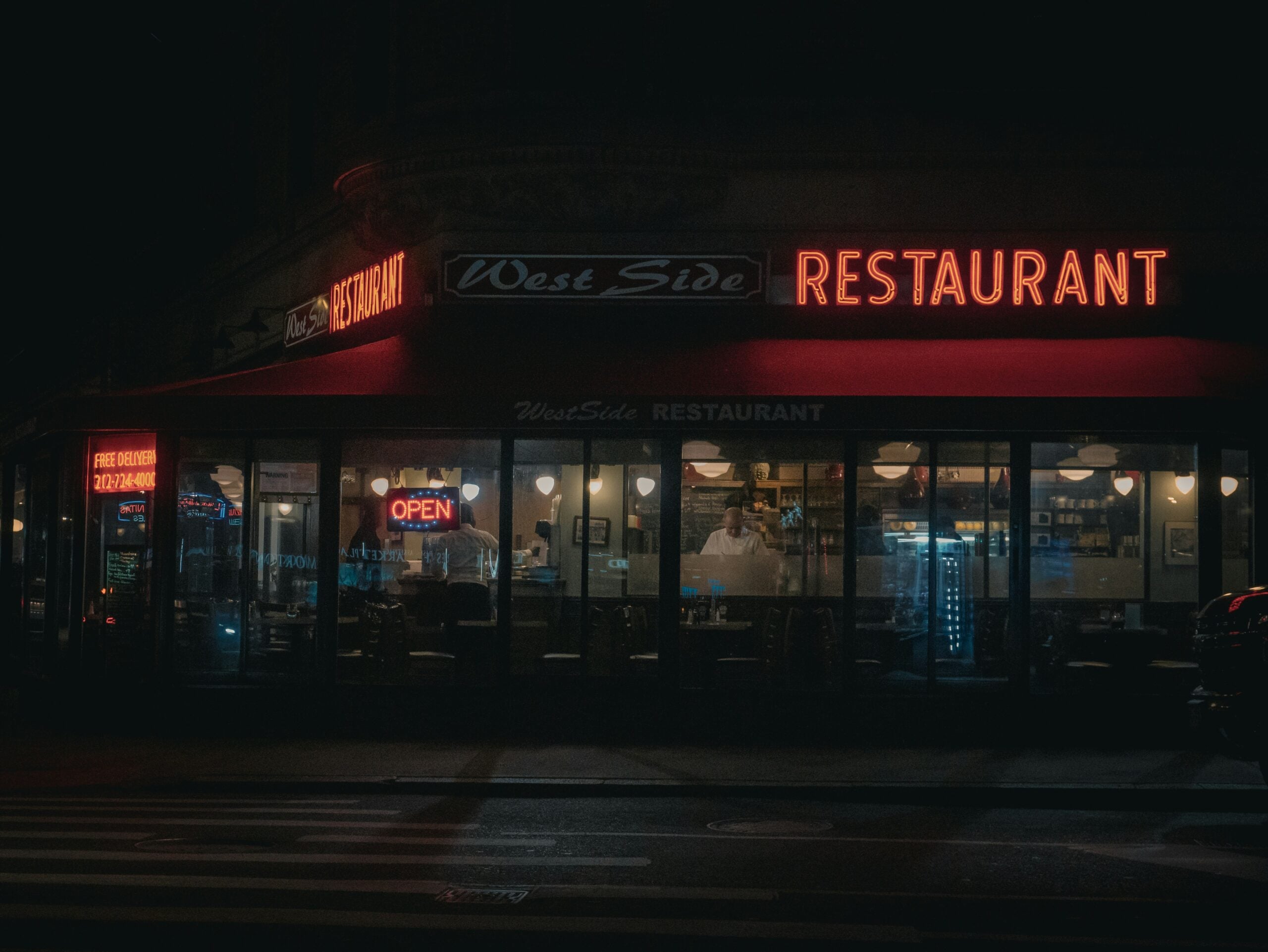 A restaurant at night with neon signs reading "West Side RESTAURANT" and "OPEN" in the windows. Warm lights illuminate the interior, showing tables, chairs, and a counter, while the street outside is quiet and dark.