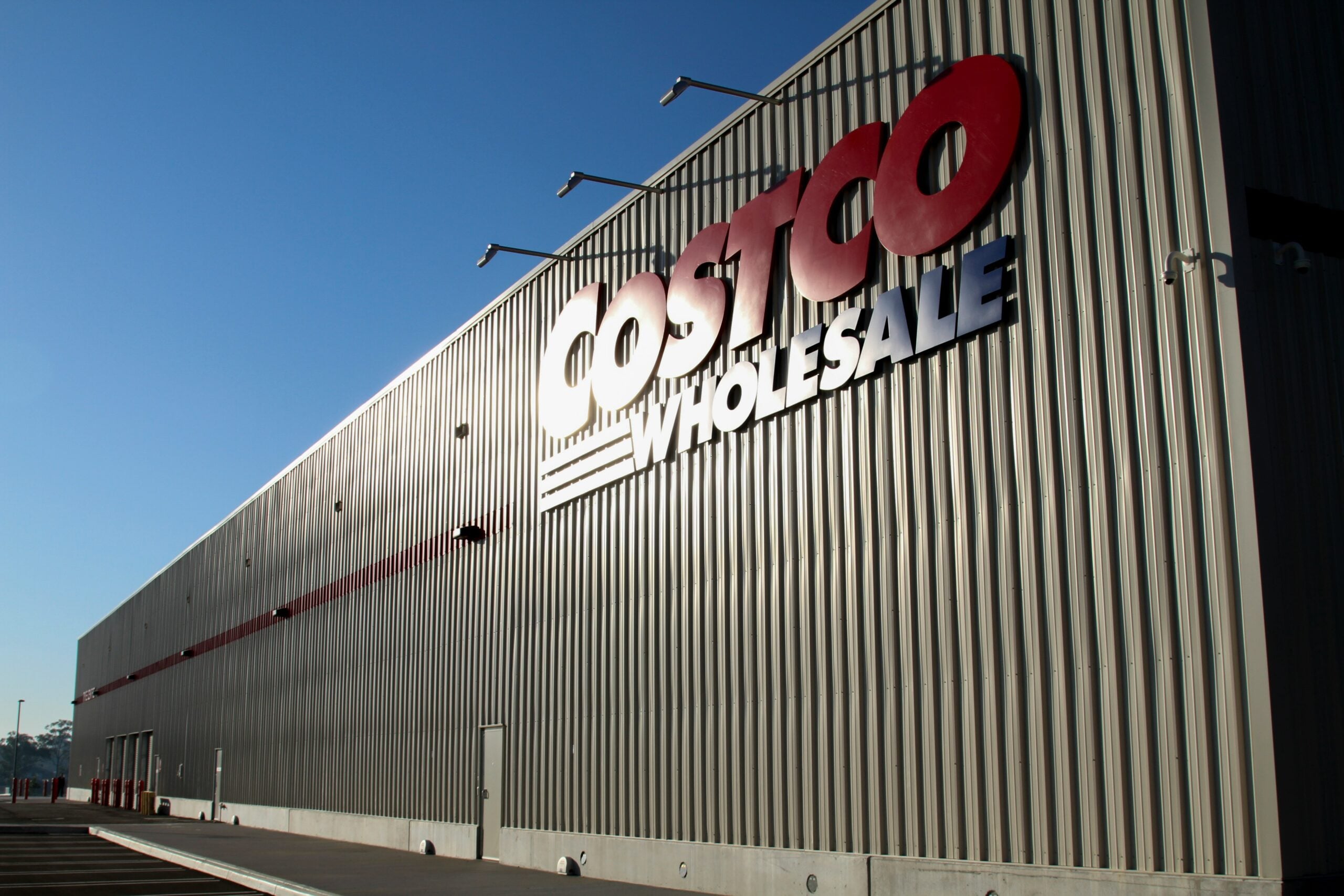 A large Costco Wholesale warehouse with a gray exterior and bold red and white signage, photographed from an angle under a clear blue sky.