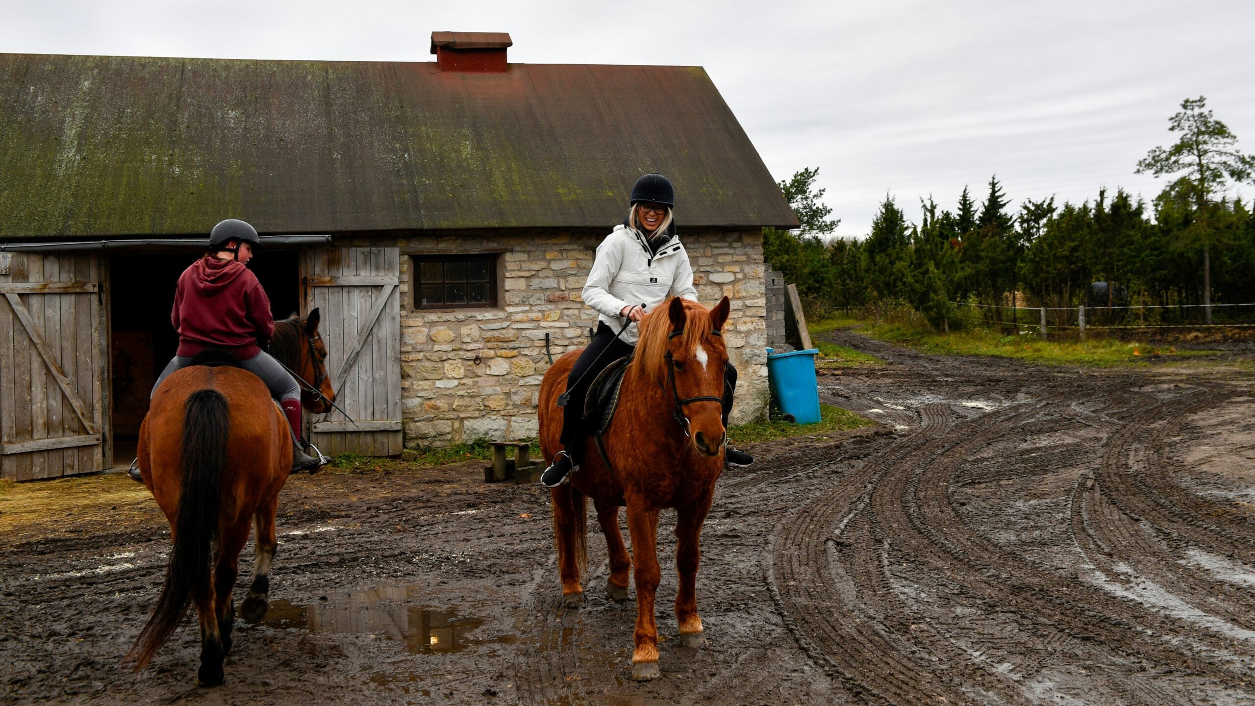 Two people wearing helmets ride horses on a muddy path near a stone barn with open wooden doors. One rider faces the camera and smiles, while the other rides away. The sky is overcast.