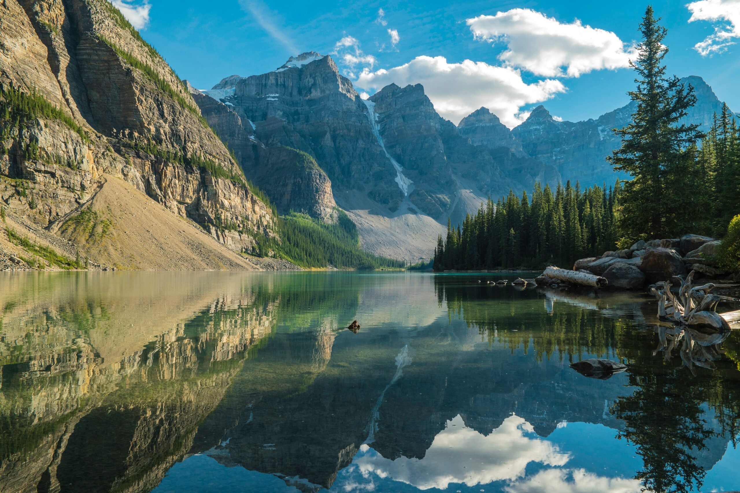 A clear mountain lake reflects rugged peaks, green pine trees, and a bright blue sky with scattered clouds; rocky shores and forested slopes frame the serene landscape.