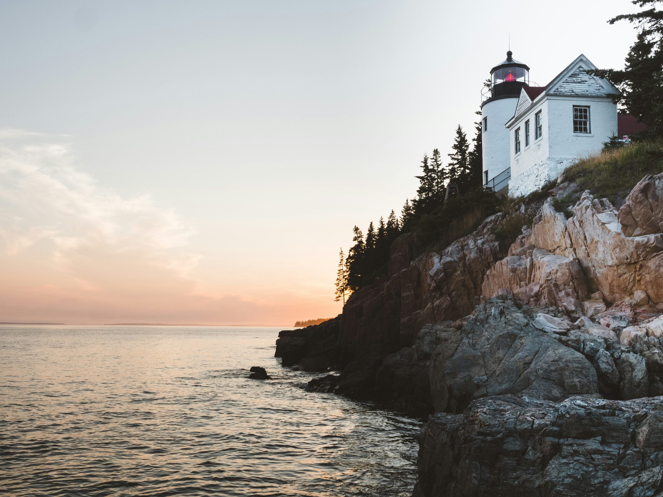 A white lighthouse with a red light stands on a rocky cliff above the ocean at sunset, surrounded by pine trees. The sky is soft with pastel colors, and gentle waves lap at the rocks below.